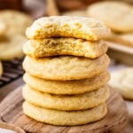 A close-up image of a stack of sugar cookies, with the top cookie partially bitten to reveal a soft, tender, and crumbly inside. The cookies have a golden-brown edge and a lighter center, showcasing their chewy texture. They are stacked on a wooden surface, with the focus on their delicious interior and perfect bake. The background is softly blurred, adding to the warm and inviting atmosphere, and a small wooden bowl of sugar is visible in the foreground, emphasizing the homemade feel of these classic cookies.