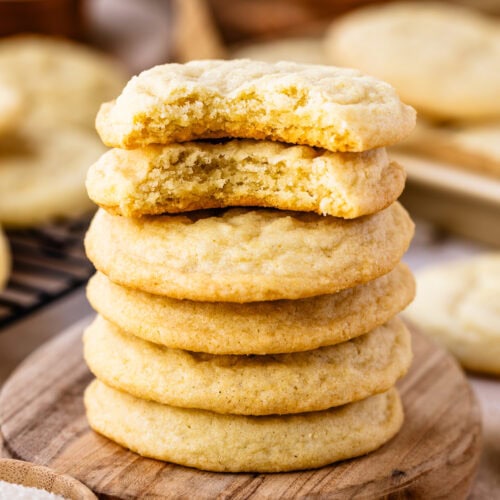 A close-up image of a stack of sugar cookies, with the top cookie partially bitten to reveal a soft, tender, and crumbly inside. The cookies have a golden-brown edge and a lighter center, showcasing their chewy texture. They are stacked on a wooden surface, with the focus on their delicious interior and perfect bake. The background is softly blurred, adding to the warm and inviting atmosphere, and a small wooden bowl of sugar is visible in the foreground, emphasizing the homemade feel of these classic cookies.