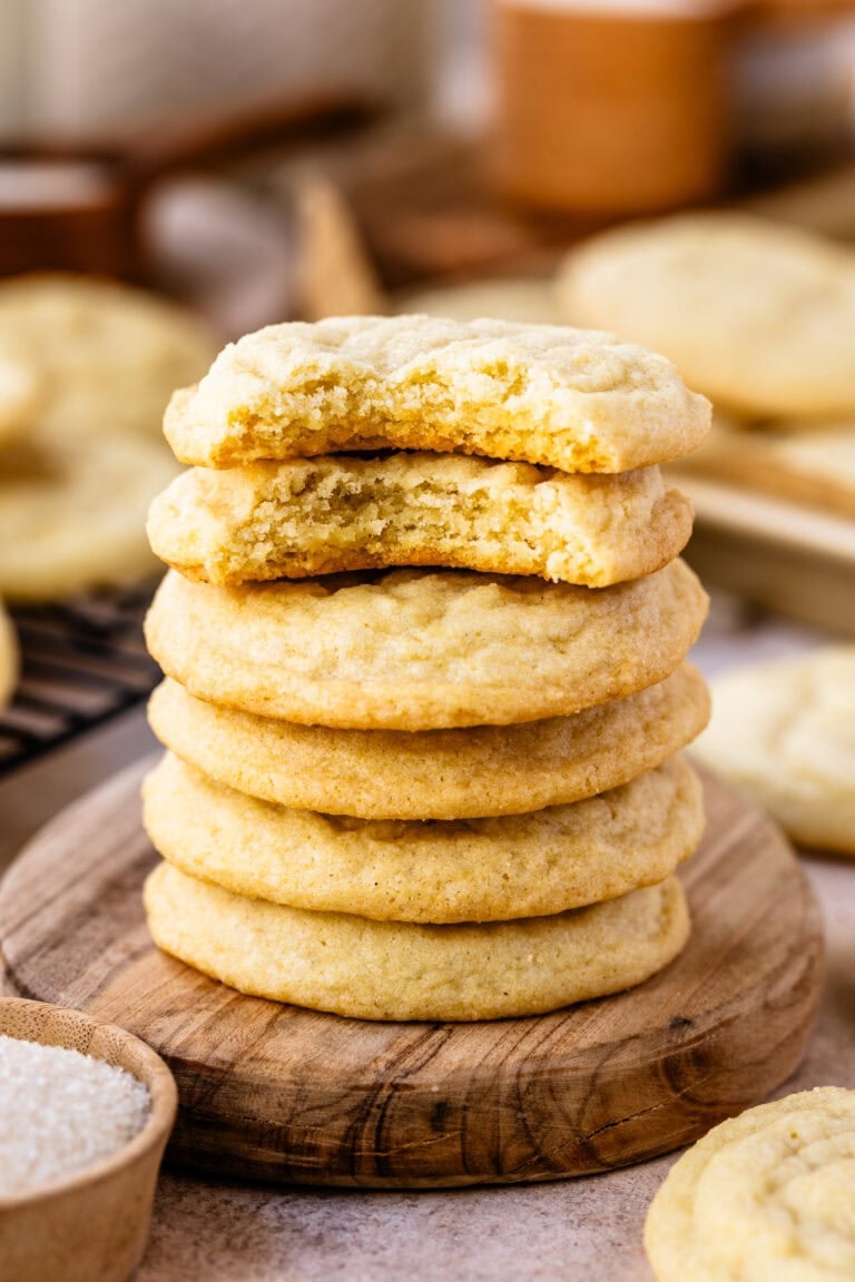 A close-up image of a stack of sugar cookies, with the top cookie partially bitten to reveal a soft, tender, and crumbly inside. The cookies have a golden-brown edge and a lighter center, showcasing their chewy texture. They are stacked on a wooden surface, with the focus on their delicious interior and perfect bake. The background is softly blurred, adding to the warm and inviting atmosphere, and a small wooden bowl of sugar is visible in the foreground, emphasizing the homemade feel of these classic cookies.