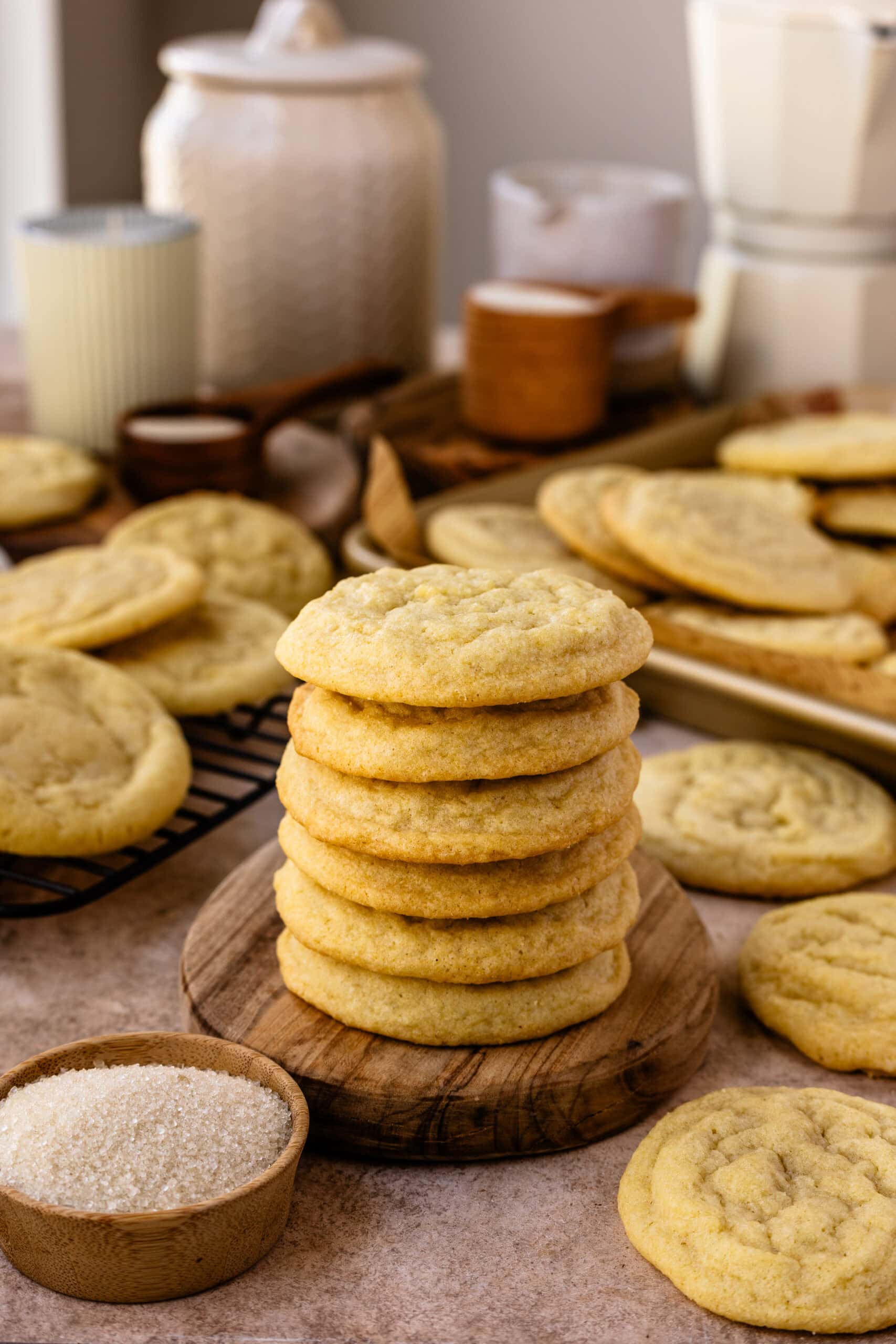 A close-up image of a stack of freshly baked sugar cookies placed on a wooden surface, showcasing their soft and slightly cracked texture. The cookies are golden brown and neatly piled to highlight their thickness and chewy nature. Surrounding the stack are more cookies scattered on a cooling rack and baking tray, with a small wooden bowl filled with sugar in the foreground. The background is softly blurred, featuring decorative items like jars, candles, and wooden utensils, contributing to the warm, cozy, and inviting atmosphere of homemade baking.