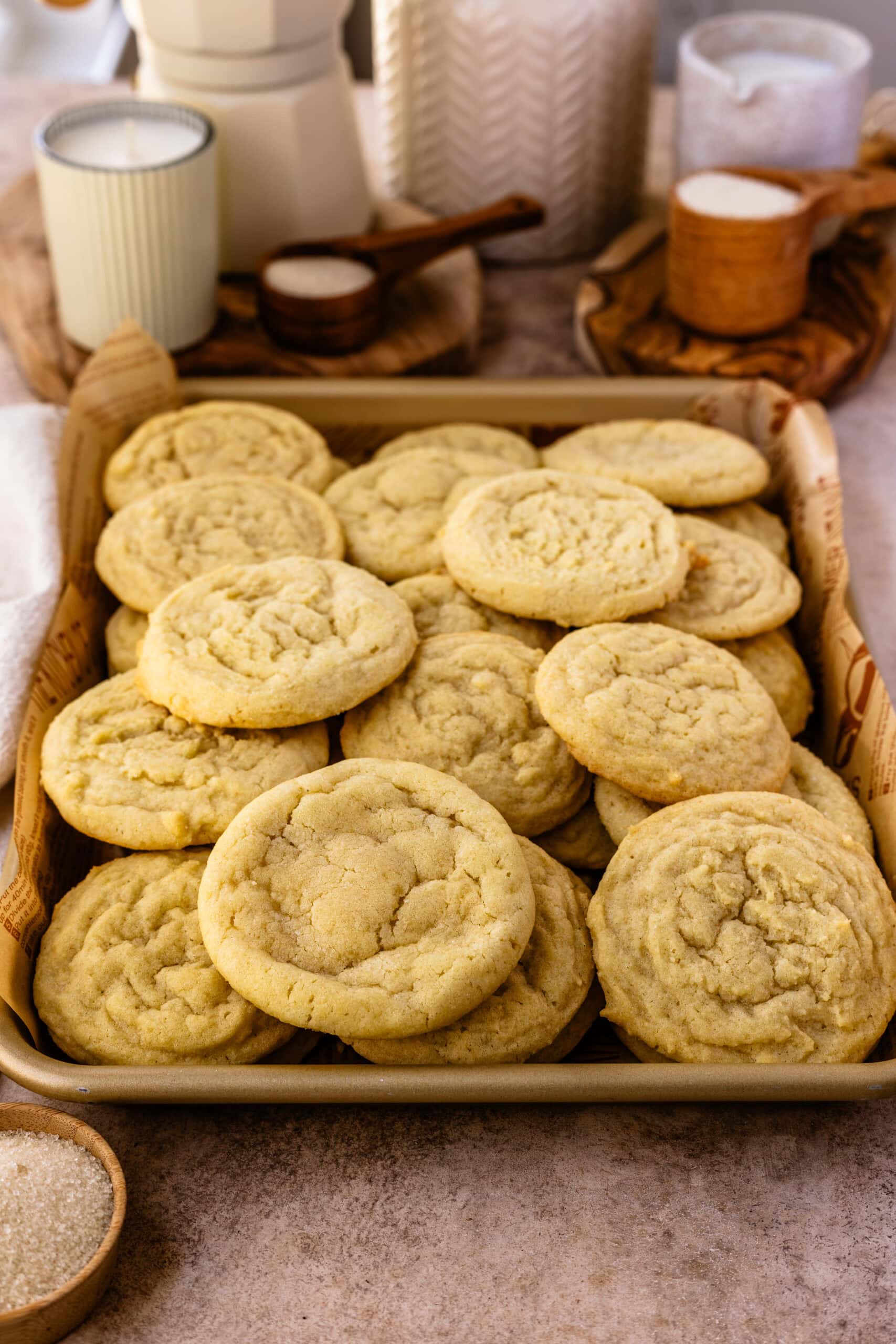 A close-up image of freshly baked sugar cookies piled onto a parchment-lined baking tray. The cookies are soft with a slightly cracked surface and a light golden-brown color, highlighting their chewy texture. The background features cozy elements like candles, wooden bowls filled with sugar, and decorative jars, enhancing the warm, inviting, and rustic feel of a home-baking environment. The cookies are presented in an appealing way, showcasing their homemade simplicity and charm.