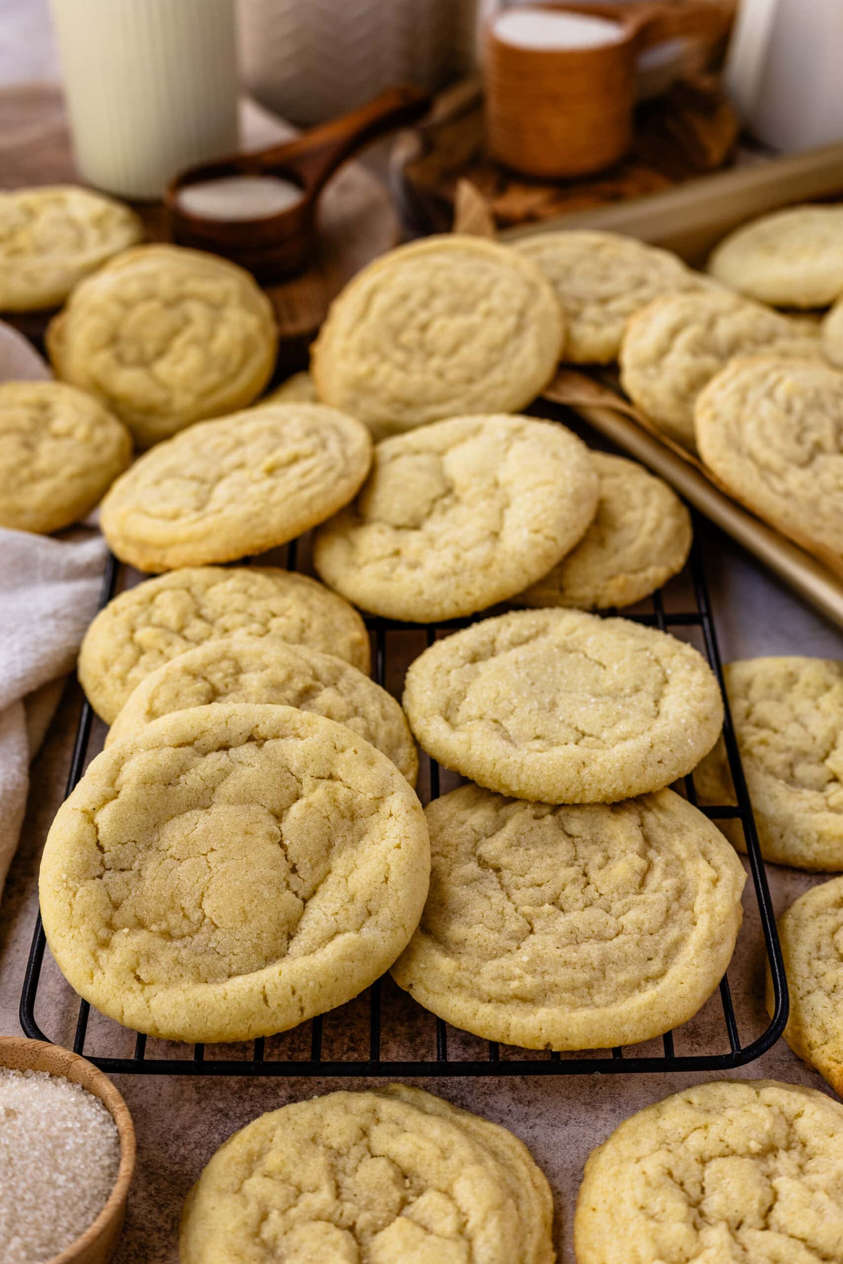 A close-up image of freshly baked sugar cookies piled on top of each other, some resting on a cooling rack. The cookies are golden with a slightly cracked surface, indicating a soft and chewy texture. In the background, there are wooden bowls filled with sugar and decorative elements like candles and jars, enhancing the warm and inviting feel of the baking scene. The image showcases the cookies beautifully, emphasizing their homemade look and classic appeal.