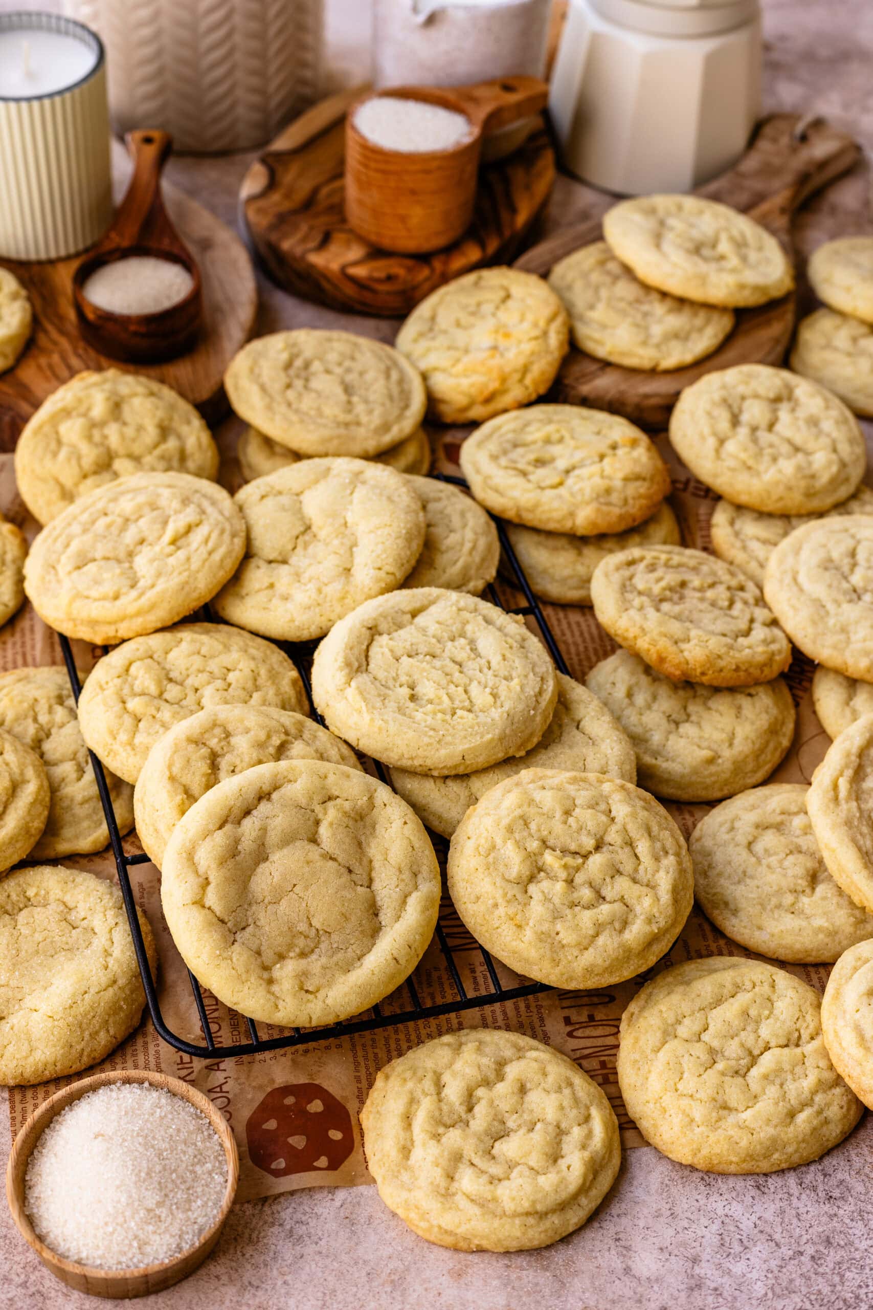 A close-up image of freshly baked sugar cookies piled on top of each other, some resting on a cooling rack. The cookies are golden with a slightly cracked surface, indicating a soft and chewy texture. In the background, there are wooden bowls filled with sugar and decorative elements like candles and jars, enhancing the warm and inviting feel of the baking scene. The image showcases the cookies beautifully, emphasizing their homemade look and classic appeal.