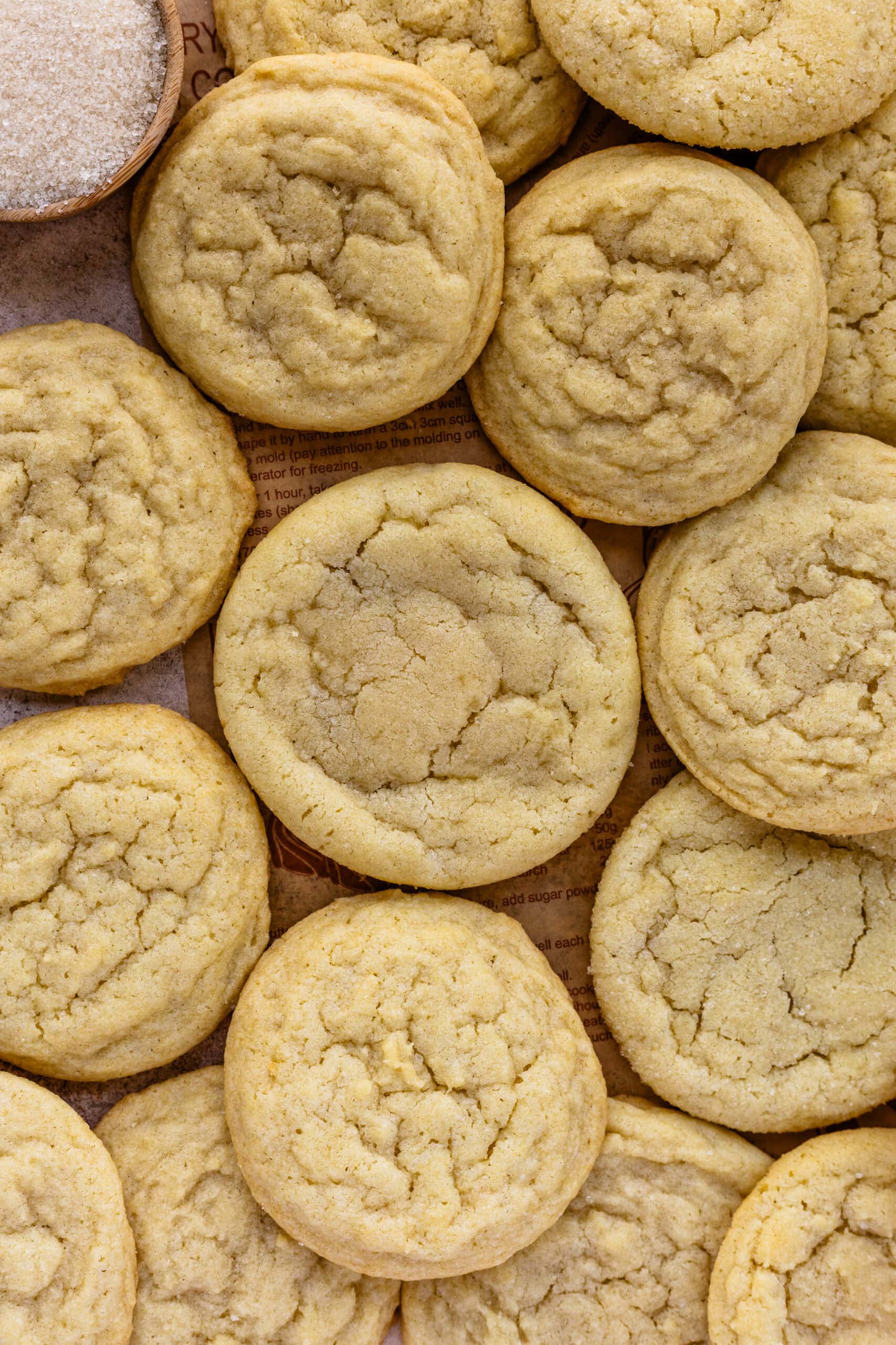 An overhead image of freshly baked sugar cookies arranged closely together, showcasing their soft and lightly cracked tops. The cookies have a golden hue and appear chewy and tender, with a slightly crisp edge. A small wooden bowl filled with sugar is visible in the corner, adding to the texture and baking ambiance. The cookies are laid out on parchment paper, creating a cozy and homemade feel that highlights their simplicity and classic appeal.