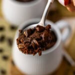 A close-up image of a spoonful of chocolate mug cake topped with melted chocolate chips. The cake has a moist, rich texture with gooey, melted chocolate chips adding extra decadence. The mug from which the cake was scooped is visible in the background, creating a warm and inviting dessert scene. The blurred background enhances focus on the indulgent spoonful, perfect for chocolate lovers.