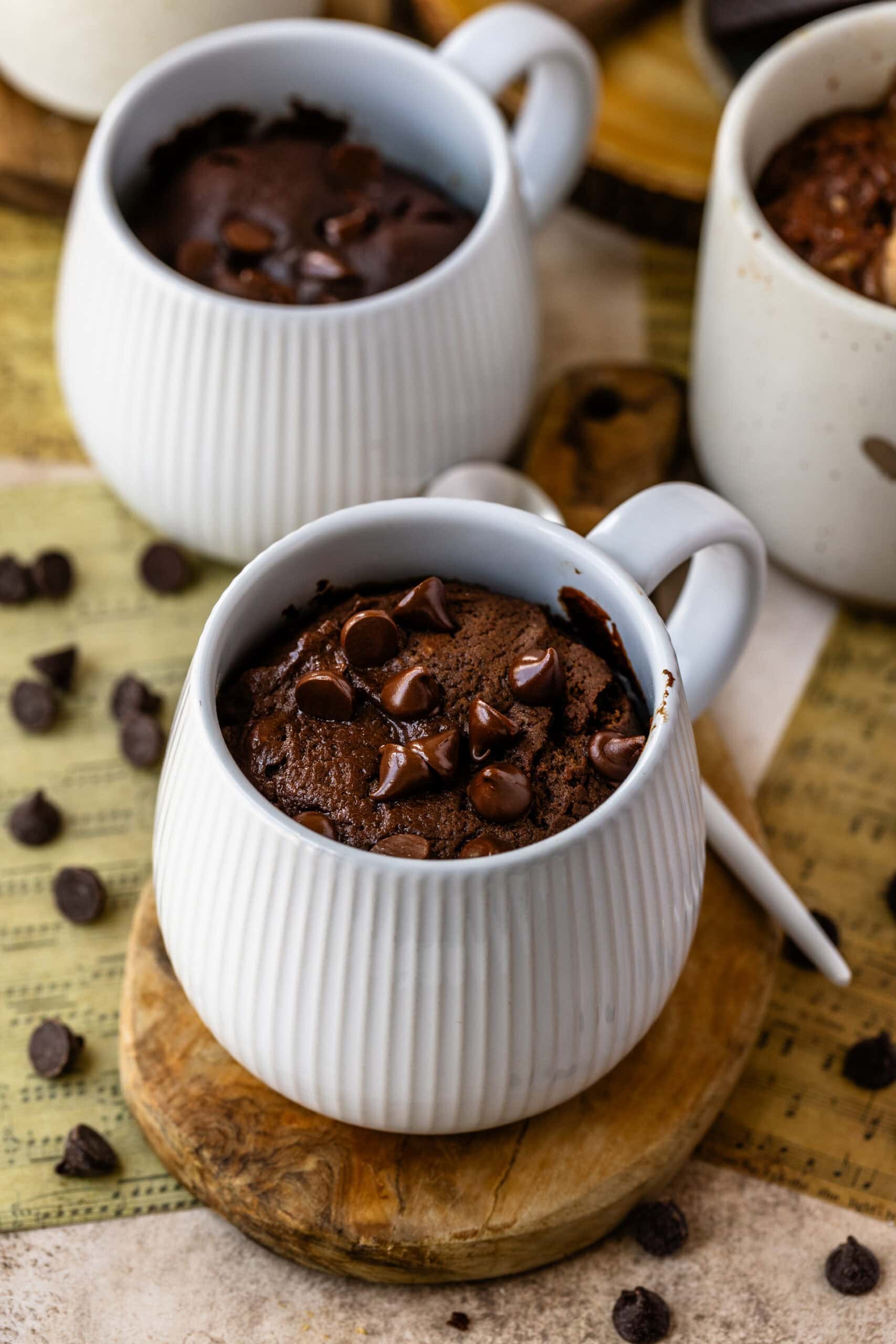 A close-up image of a chocolate mug cake topped with melted chocolate chips, served in a white ribbed ceramic mug. The cake is fluffy and rich, with gooey chocolate pockets visible on the surface. The mug sits on a small wooden board, with scattered chocolate chips around for decoration, giving the setup a warm and cozy feel.
