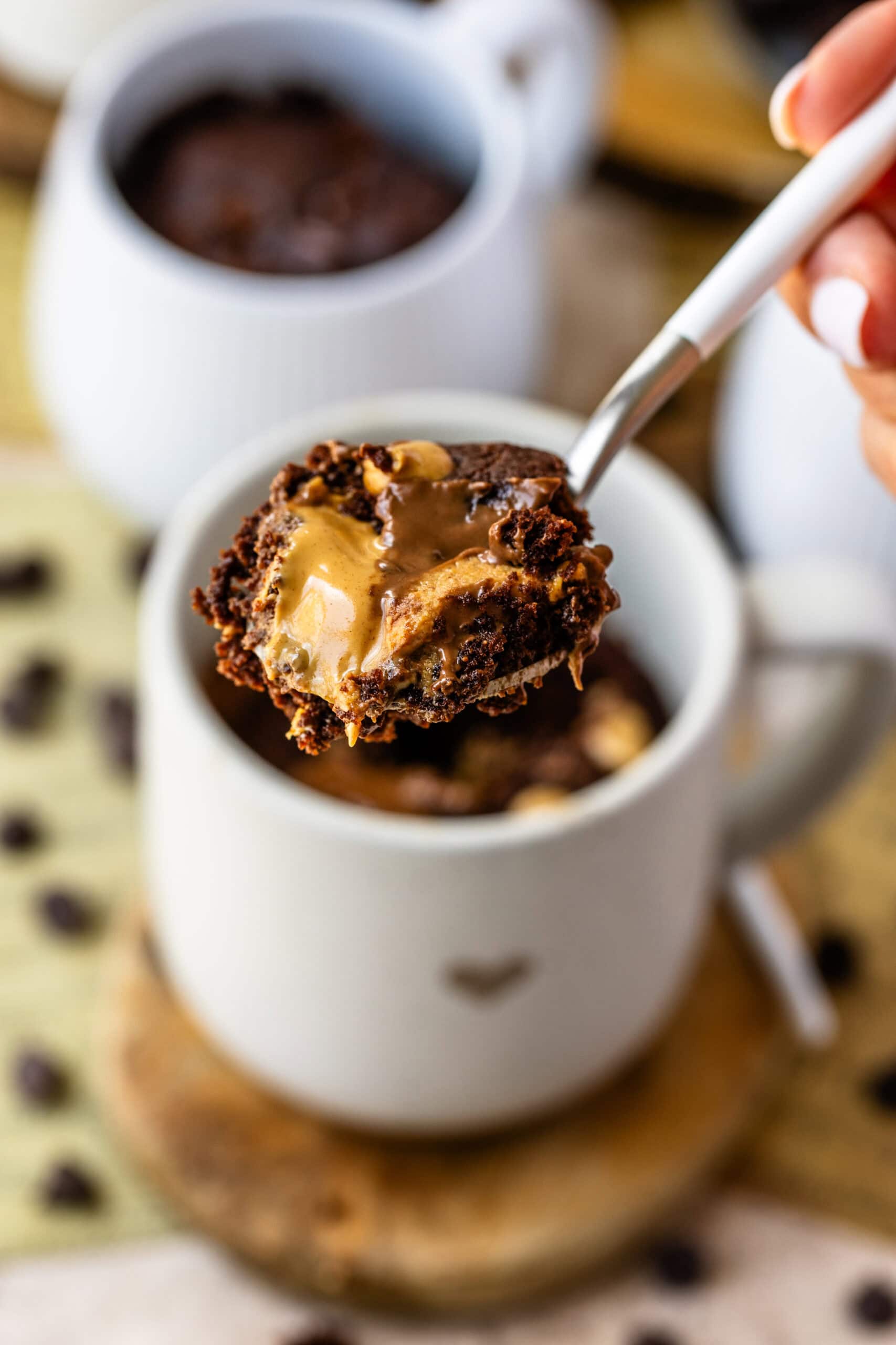 A close-up image of a spoonful of chocolate mug cake with a gooey peanut butter center. The rich, moist cake texture contrasts with the creamy peanut butter swirl, creating a decadent bite. The mug with the remaining cake sits below on a small wooden board, with a subtle heart design visible on the mug. Blurred in the background are other mugs, enhancing the cozy, inviting feel of the image.