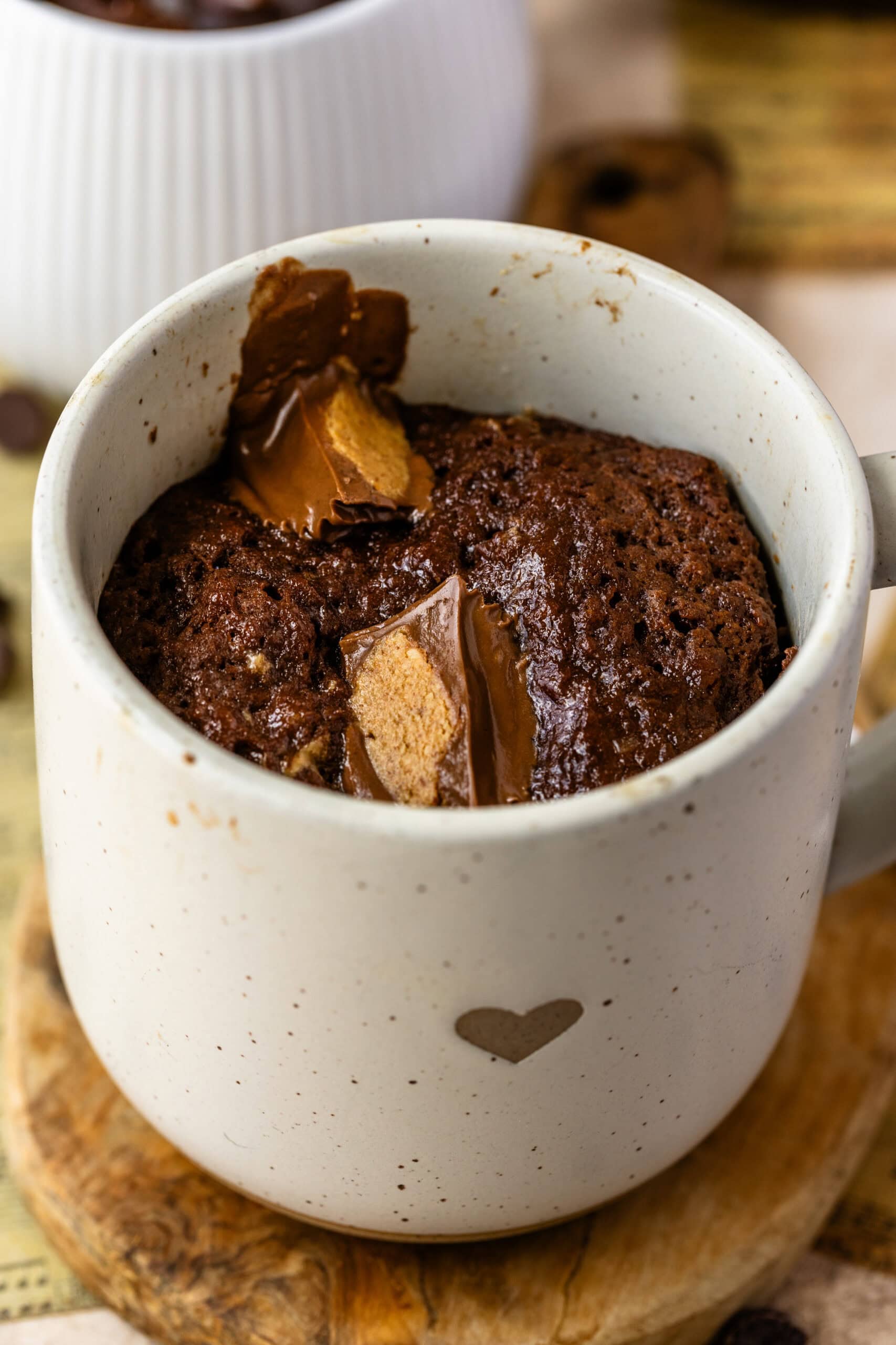 A close-up image of a freshly baked chocolate mug cake with chunks of peanut butter cup melted on top. The cake is rich and moist, with the peanut butter cup adding a creamy, nutty texture. The mug has a small heart design, enhancing the warm, cozy presentation. The cake sits on a wooden board, with another mug blurred in the background, giving a comforting, inviting feel to the setup.