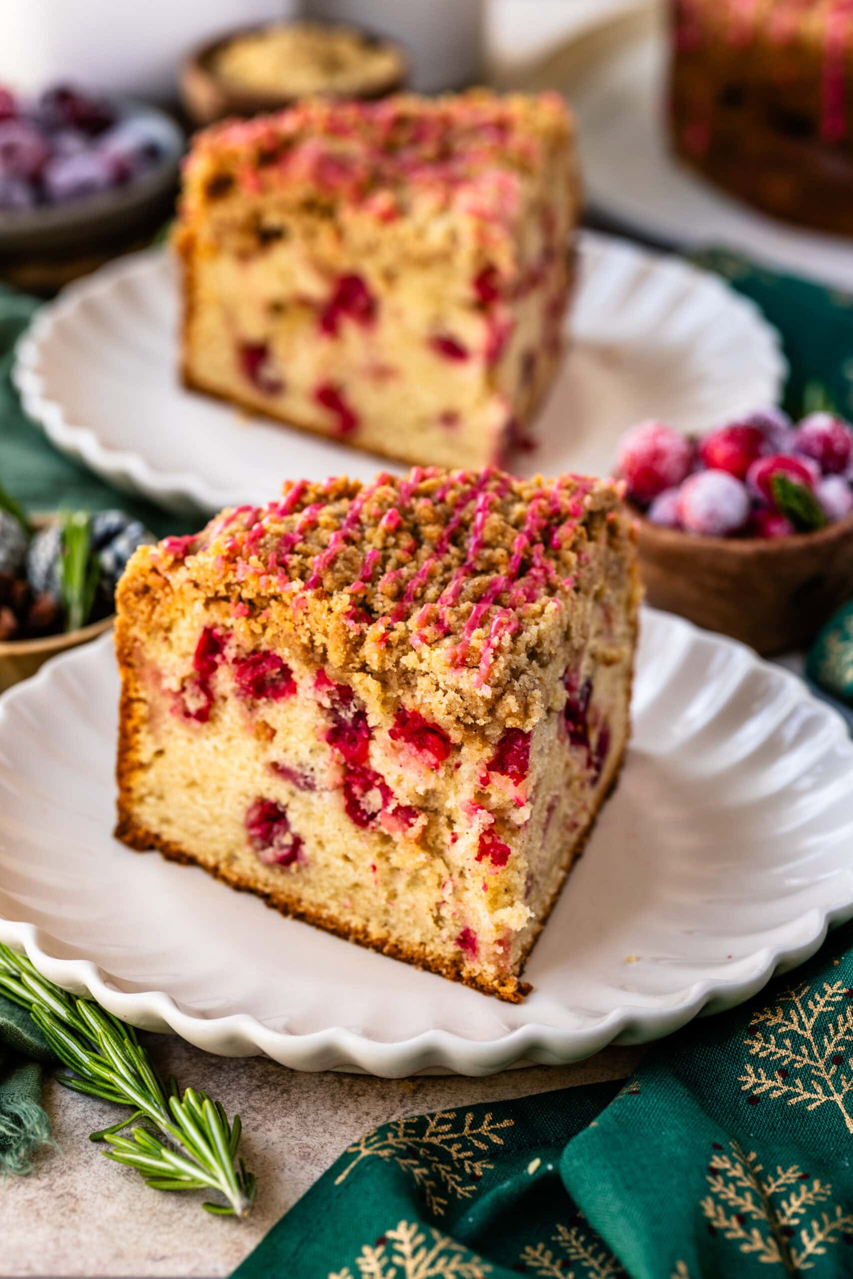 A close-up image of a slice of cranberry coffee cake on a white scalloped plate. The cake has a golden-brown crumb topping drizzled with a pink cranberry glaze, and bright red cranberries are visible throughout the moist cake. In the background, another slice is visible, along with sugared cranberries, fresh rosemary sprigs, and festive holiday decor, adding a cozy and seasonal atmosphere to the scene.