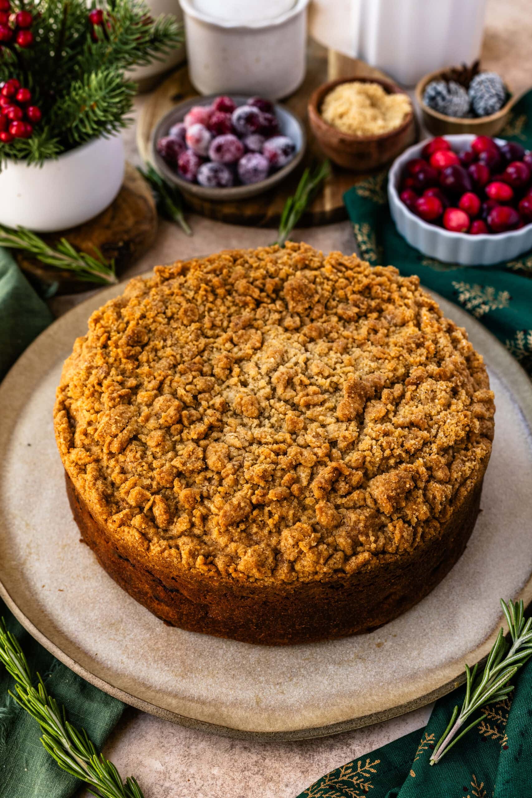 A whole cranberry coffee cake with a golden-brown crumb topping, presented on a rustic plate. The crumb layer appears perfectly baked, with a crunchy texture that contrasts the soft cake underneath. The background features holiday elements such as fresh and sugared cranberries, brown sugar, and pine cones, along with sprigs of rosemary and a green napkin, adding a festive and cozy touch to the scene.