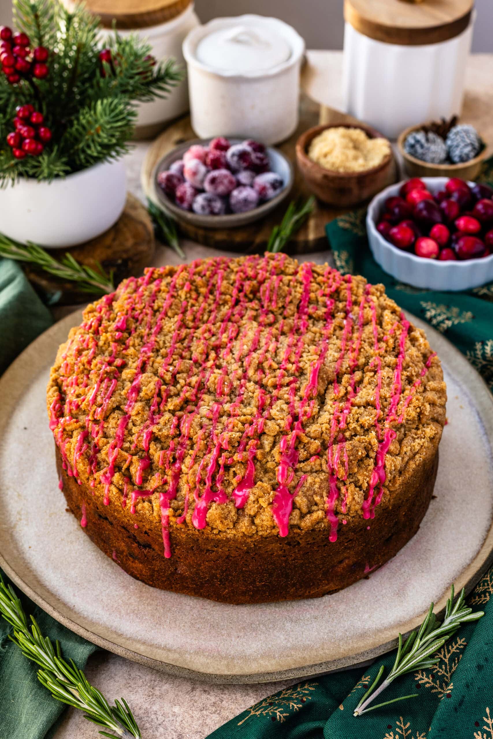 A whole cranberry coffee cake displayed on a rustic plate, topped with a golden-brown crumb layer and drizzled with a bright pink cranberry glaze. The background includes festive elements like fresh and sugared cranberries, brown sugar, and pine cones, creating a cozy, holiday-inspired scene. Fresh rosemary sprigs and a green napkin with gold details enhance the seasonal feel.