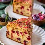 A close-up image of a slice of cranberry coffee cake on a white scalloped plate. The cake has a golden-brown crumb topping drizzled with a pink cranberry glaze, and bright red cranberries are visible throughout the moist cake. In the background, another slice is visible, along with sugared cranberries, fresh rosemary sprigs, and festive holiday decor, adding a cozy and seasonal atmosphere to the scene.
