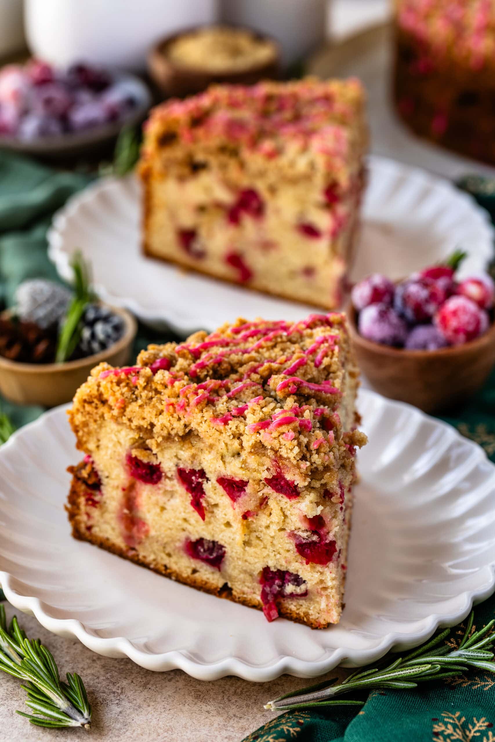A close-up image of a slice of cranberry coffee cake on a white scalloped plate. The cake has a golden-brown crumb topping drizzled with a pink cranberry glaze, and bright red cranberries are visible throughout the moist cake. In the background, another slice is visible, along with sugared cranberries, fresh rosemary sprigs, and festive holiday decor, adding a cozy and seasonal atmosphere to the scene.