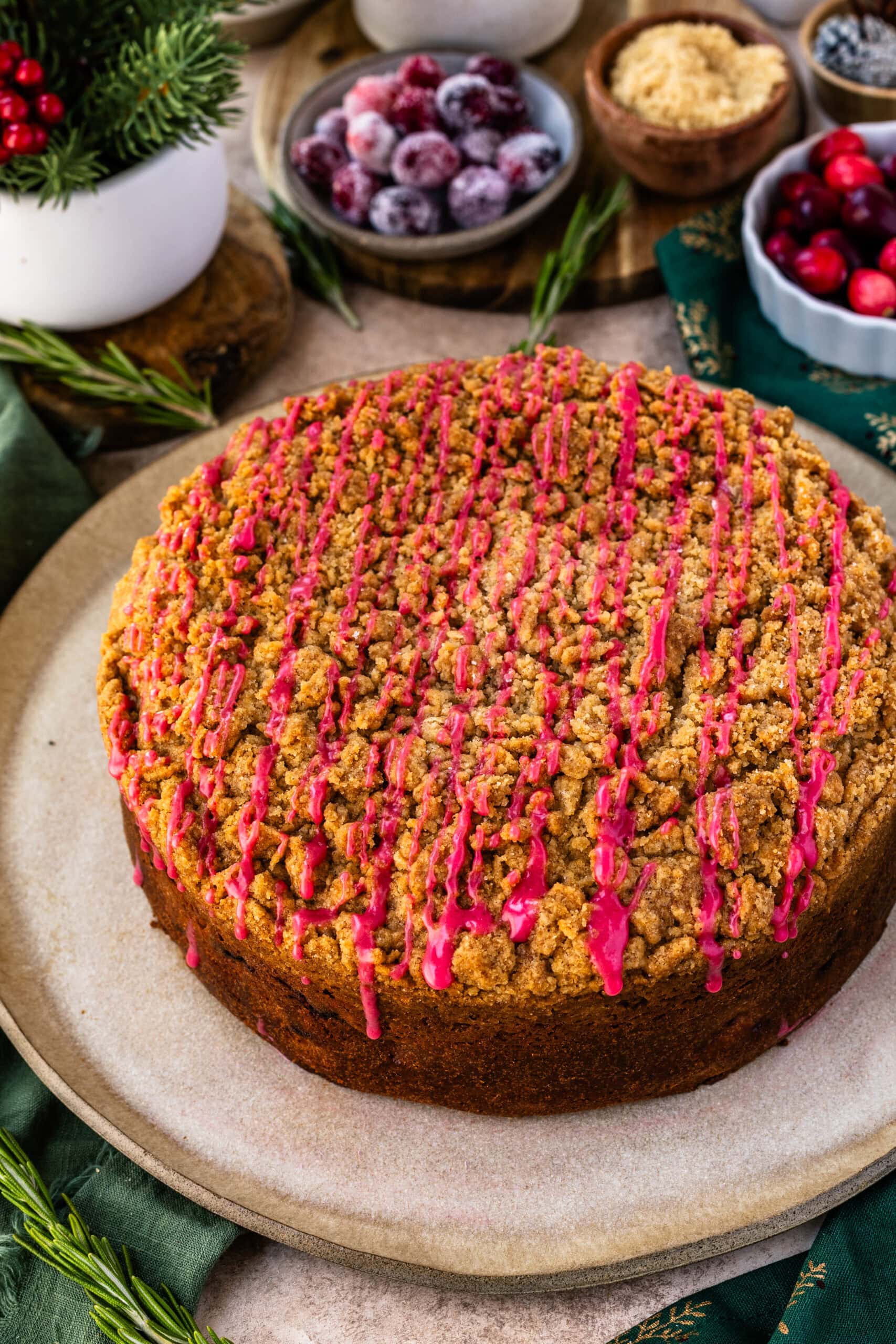A whole cranberry coffee cake displayed on a rustic plate, topped with a golden-brown crumb layer and drizzled with a bright pink cranberry glaze. The background includes festive elements like fresh and sugared cranberries, brown sugar, and pine cones, creating a cozy, holiday-inspired scene. Fresh rosemary sprigs and a green napkin with gold details enhance the seasonal feel.