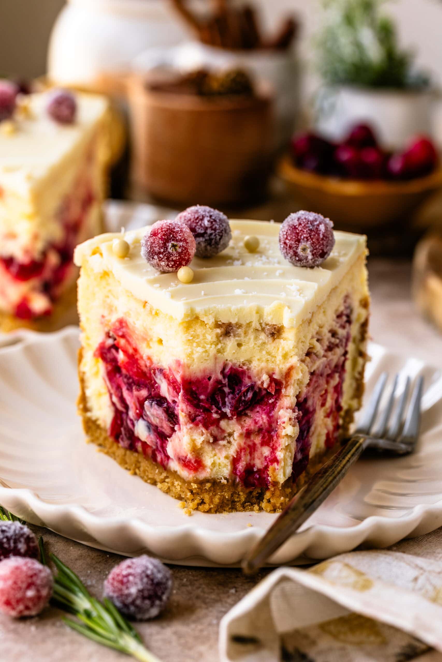 A close-up shot of a square slice of cranberry cheesecake on a scalloped plate, showcasing its creamy texture and vibrant cranberry swirls. The top is decorated with sugared cranberries and white chocolate pearls, adding a festive touch. The graham crust at the base provides a sturdy foundation. In the background, rustic elements like fresh cranberries and greenery enhance the holiday ambiance, while additional sugared cranberries and a sprig of rosemary rest beside the plate for added elegance.