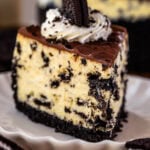 Close-up of a single slice of Oreo cheesecake on a scalloped plate, featuring an Oreo crust, creamy cheesecake swirled with Oreo pieces, chocolate ganache topping, whipped cream, and an Oreo cookie garnish, with blurred cheesecake in the background.