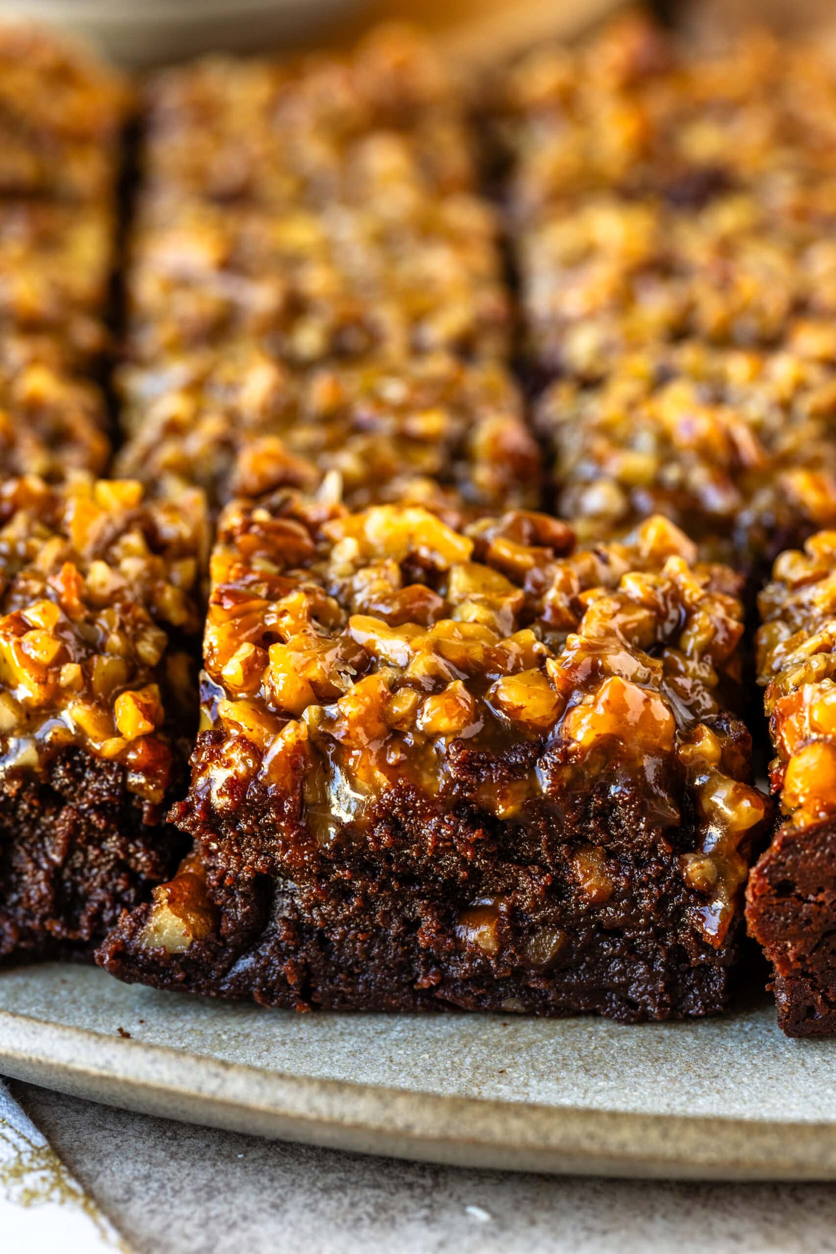 This image shows close-up pecan pie brownies, beautifully layered with a dense, fudgy brownie base and a caramelized pecan topping. The texture of the brownie is rich and moist, contrasting with the glossy, crunchy pecans that glisten under a layer of caramel. The pieces are arranged in neat rows on a rustic plate, highlighting the golden-brown topping that makes these brownies look indulgent and irresistibly delicious.