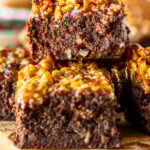 A close-up of stacked pecan pie brownies, highlighting the rich, fudgy chocolate layers and the caramelized pecan topping. The brownie on top displays a moist, dense crumb, with chopped pecans glistening in a golden caramel glaze. The layers show the perfect contrast between the gooey topping and the thick, decadent brownie base. The background is softly blurred, focusing all attention on the texture and richness of the dessert, giving it a warm, inviting look.