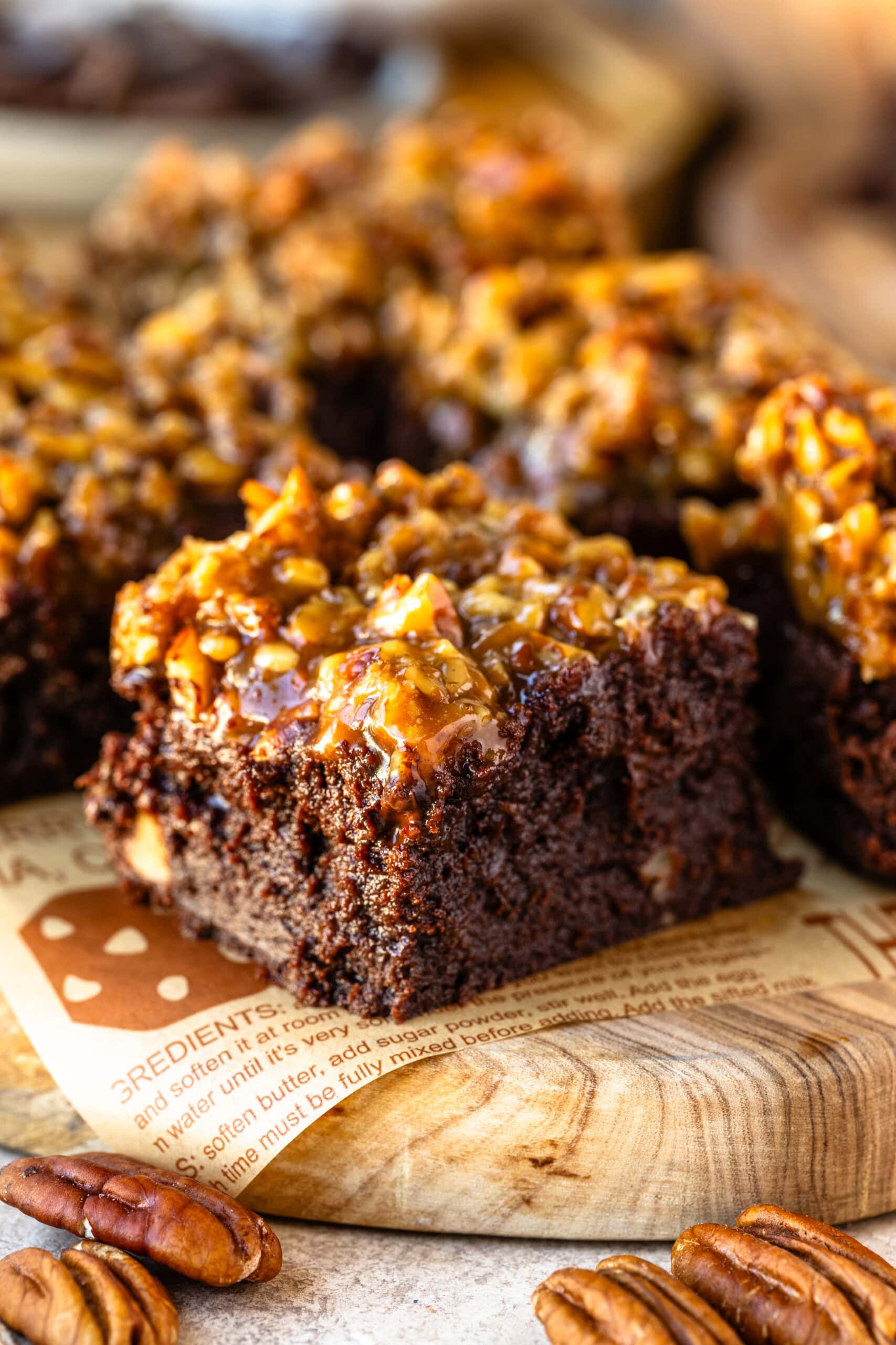 A close-up of a single pecan pie brownie on a wooden board, showcasing its dense, fudgy chocolate layer topped with a glossy, caramelized pecan mixture. The brownie has a rich texture, and the pecan topping glistens with a golden caramel coating, giving it a deliciously gooey and crunchy appearance. Whole pecans are scattered around the board, adding a rustic touch to the scene and enhancing the inviting, homemade vibe of the dessert.