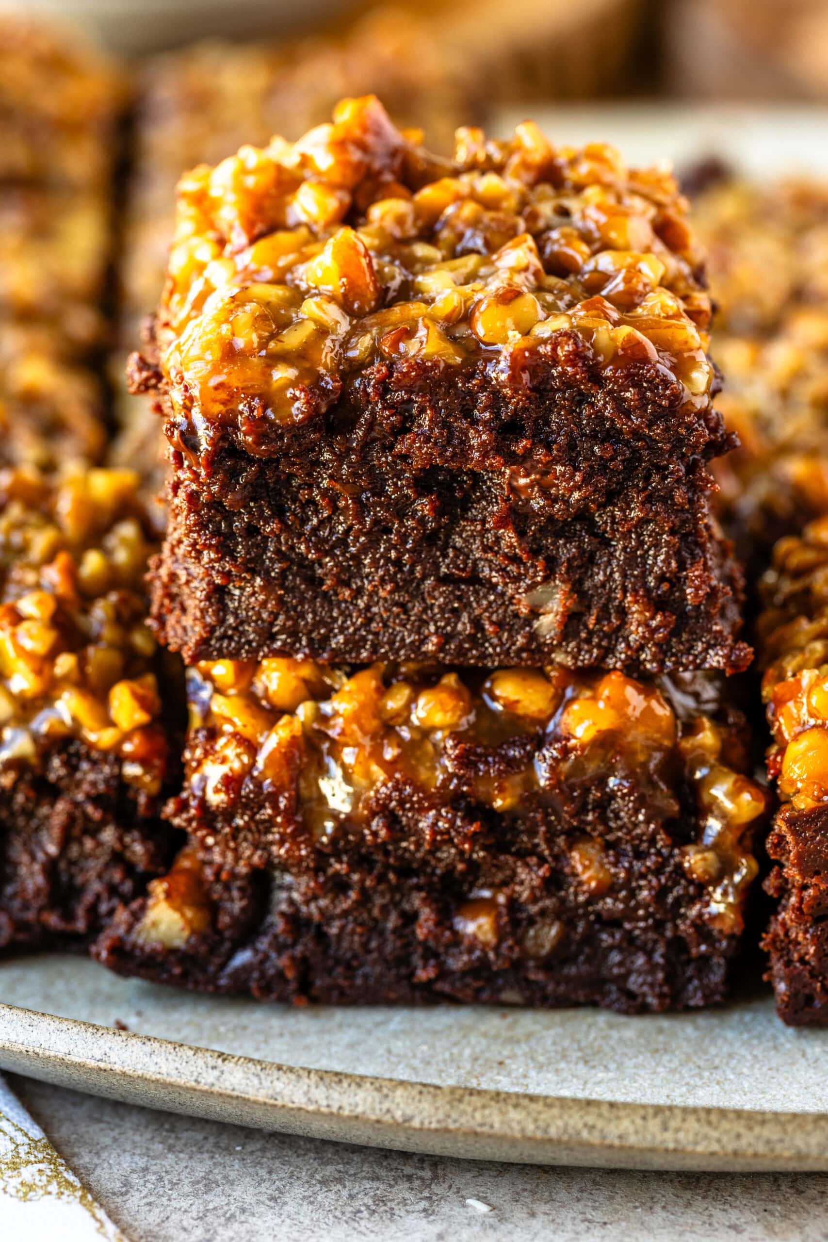 A close-up of two pecan pie brownies stacked on top of each other, showcasing their dense, fudgy chocolate base and a rich, caramelized pecan topping. The top layer glistens with a glossy, golden pecan mixture, adding a crunchy and gooey texture contrast to the soft, moist brownie beneath. The intricate details of the pecan pieces and caramel coating highlight the indulgent nature of these brownies, arranged on a rustic ceramic plate for a cozy, inviting presentation.