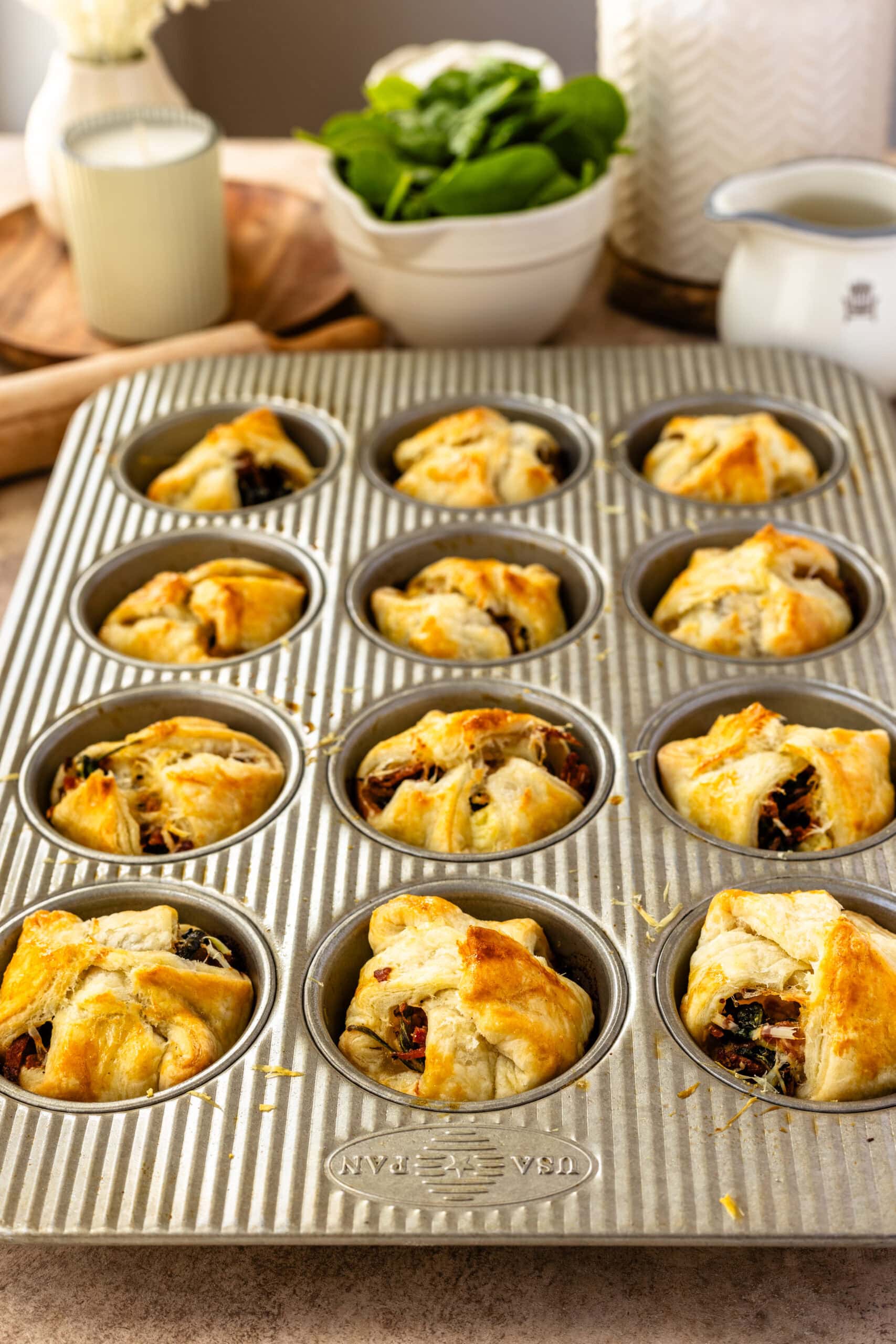 Freshly baked spinach puffs in a muffin tin, each with a golden-brown, flaky crust and savory filling peeking out slightly from the pastry folds. The background shows kitchen items like a bowl of fresh spinach, a rolling pin, and a jug, adding a homely and appetizing setting to the image.
