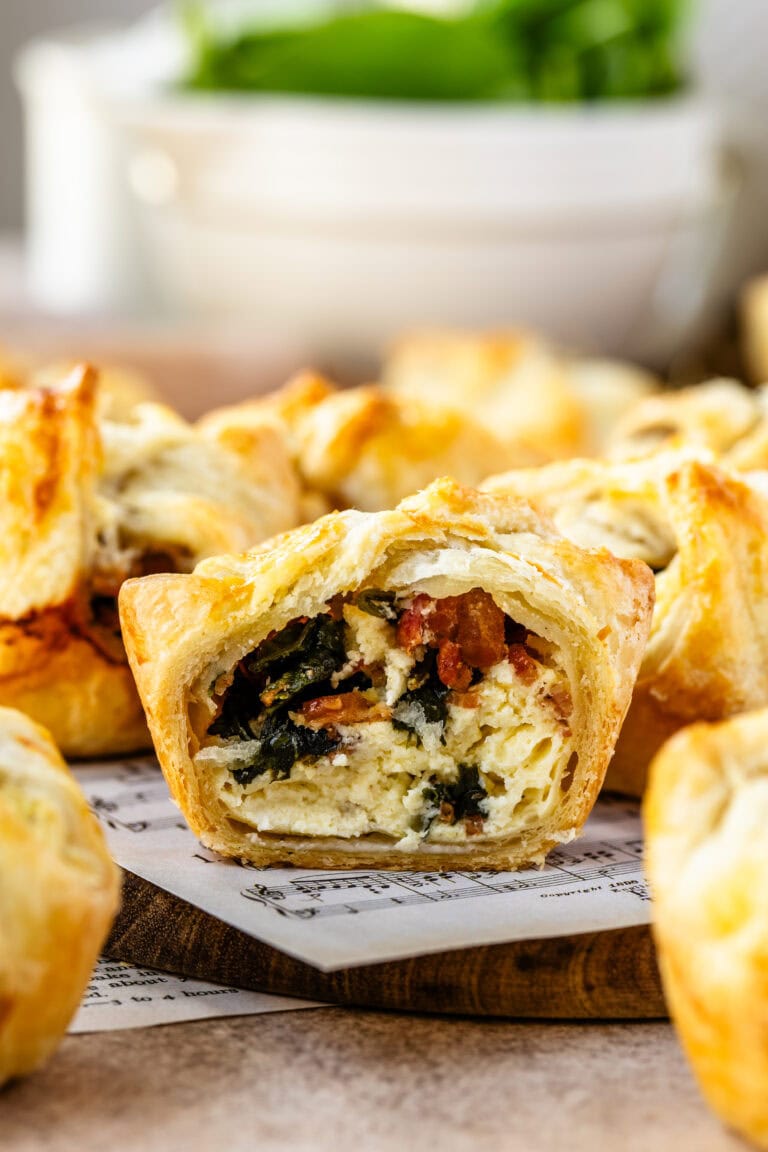 Close-up image of a spinach puff with a bite taken out, revealing the filling of creamy cheese, spinach, and bacon inside the flaky golden pastry. Other spinach puffs are in the background, showcasing the appetizing texture and layers of the pastry and filling.