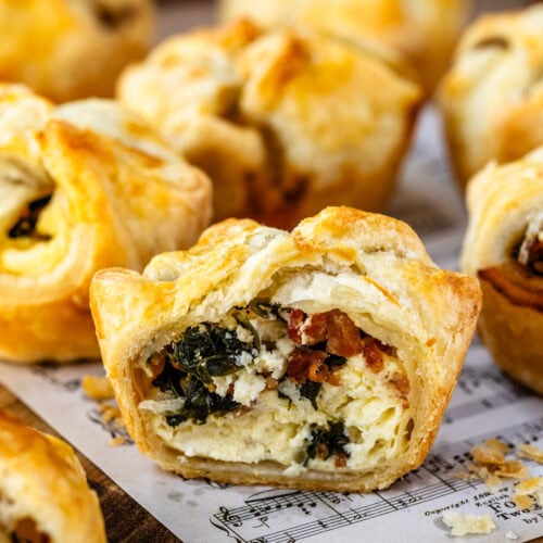 Close-up image of a spinach puff with a bite taken out, revealing the filling of creamy cheese, spinach, and bacon inside the flaky golden pastry. Other spinach puffs are in the background, showcasing the appetizing texture and layers of the pastry and filling.