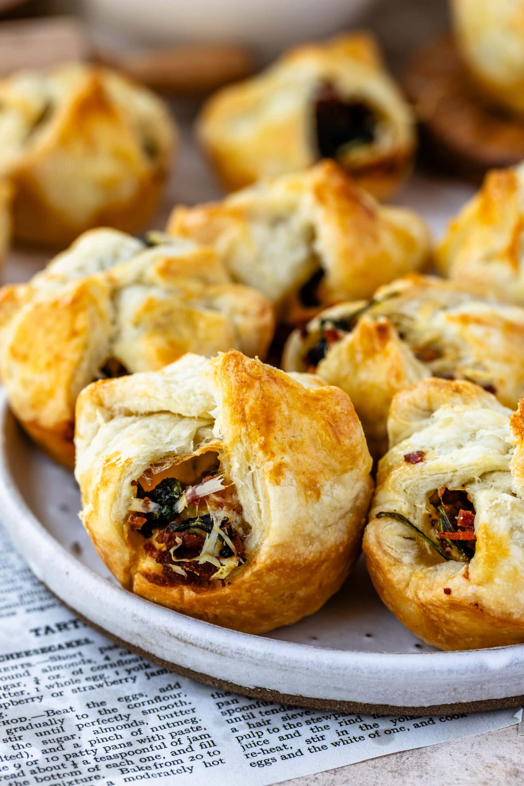 A plate of freshly baked spinach puffs with golden, flaky pastry and savory filling, arranged on a round white plate. The spinach, bacon, and cheese filling is visible in some puffs, adding a rich, appetizing texture. In the background, there are kitchen items, including a bowl of fresh spinach and a rolling pin, creating a cozy and inviting setting for these delicious appetizers.