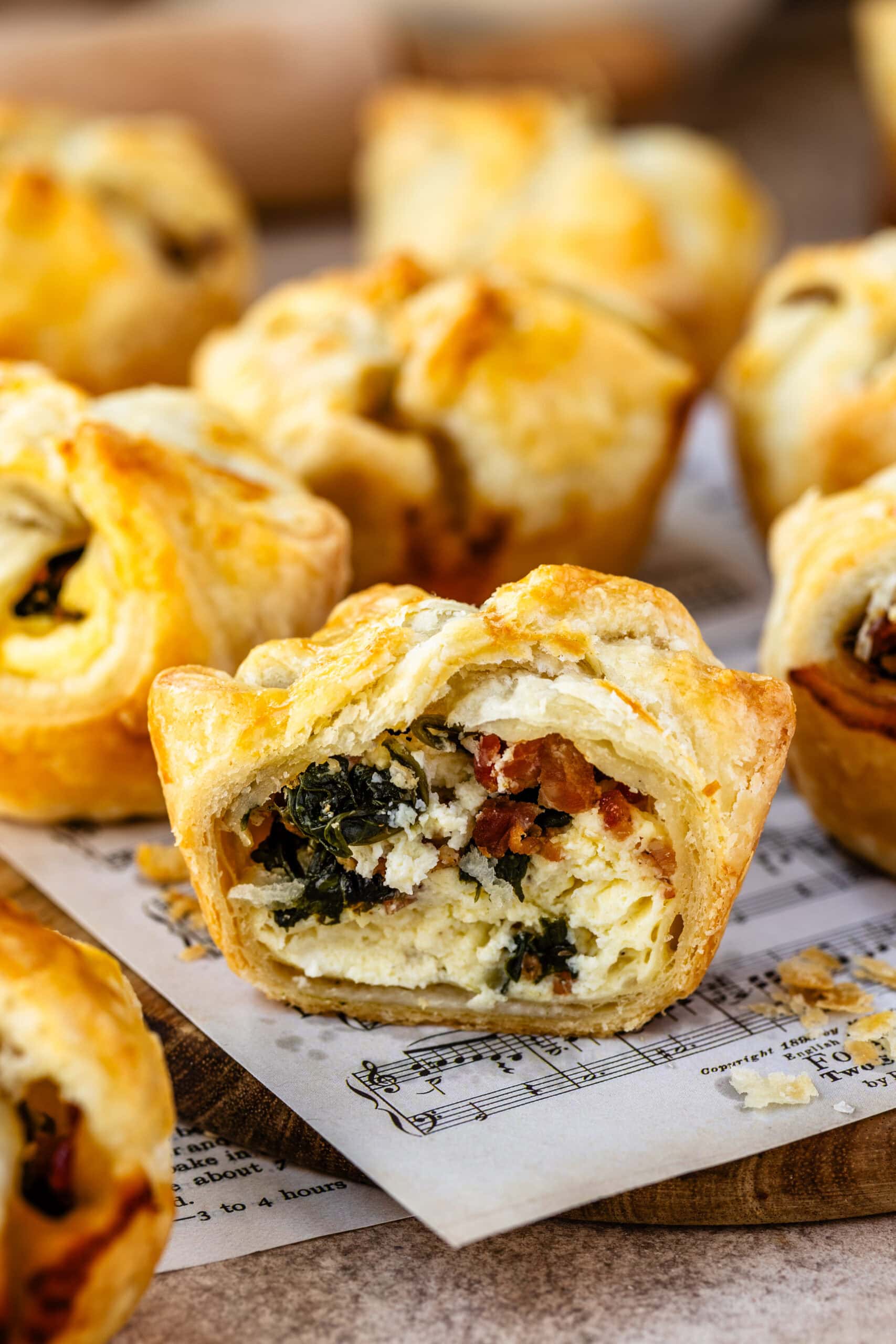 Close-up image of a spinach puff with a bite taken out, revealing the filling of creamy cheese, spinach, and bacon inside the flaky golden pastry. Other spinach puffs are in the background, showcasing the appetizing texture and layers of the pastry and filling.