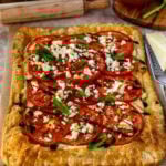 Freshly baked tomato tart with golden puff pastry crust topped with sliced tomatoes, crumbled cheese, and fresh basil leaves. The tart is set on parchment paper with scattered herbs and cooking utensils, creating a rustic presentation.