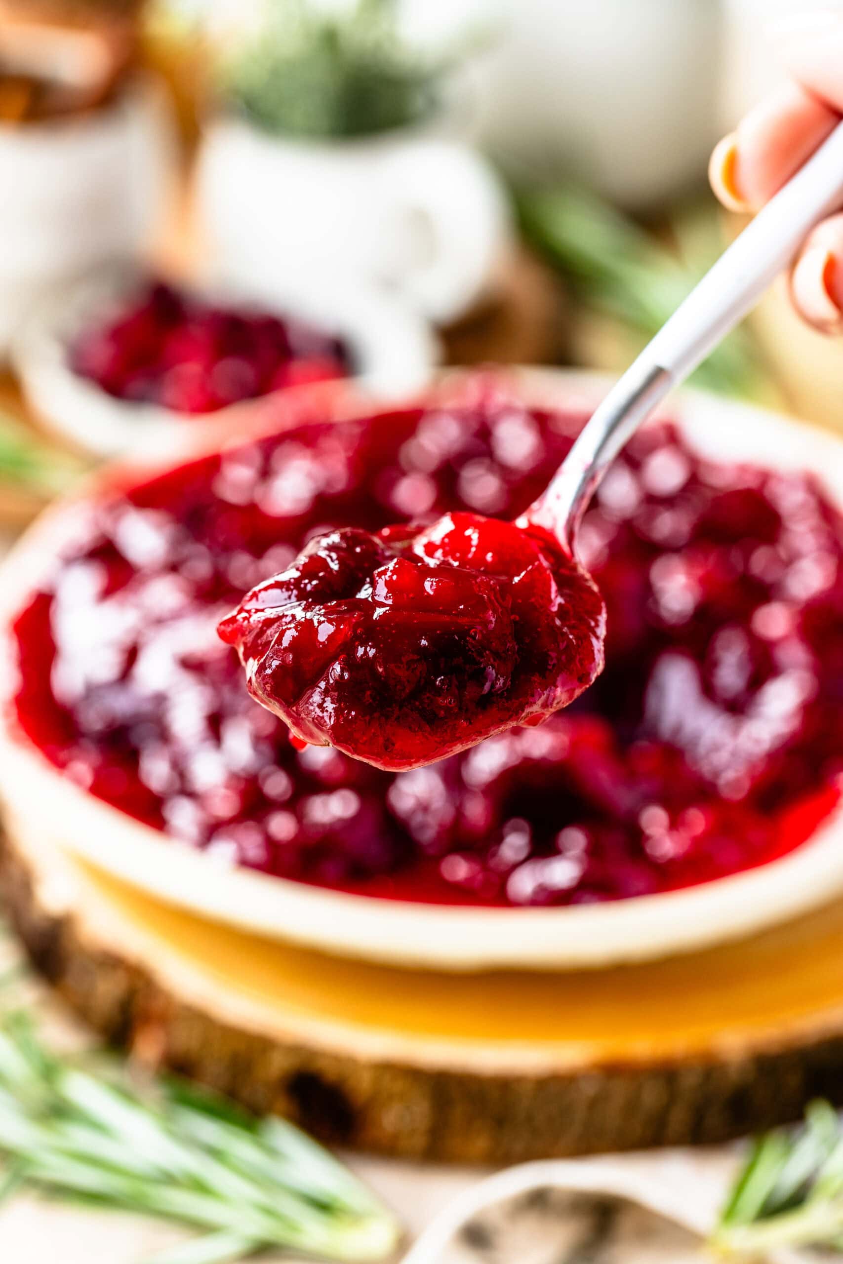 This image provides a close-up view of a spoonful of glossy cranberry sauce, showcasing its thick, chunky texture with whole cranberries and a beautiful deep red hue. The background is softly blurred, featuring a bowl filled with more cranberry sauce, set on a rustic wooden surface, adding warmth to the scene. Fresh rosemary sprigs and other seasonal decor are subtly visible, creating a festive and cozy holiday ambiance. The vibrant color and rich texture of the sauce are inviting, making it look delicious and perfect for a holiday spread.