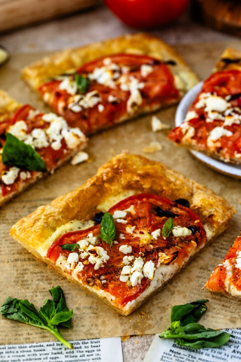 Several slices of tomato tart arranged on parchment paper, showcasing a golden puff pastry crust topped with roasted tomato slices, crumbled cheese, and fresh basil leaves. A few basil sprigs are scattered nearby, enhancing the rustic presentation.