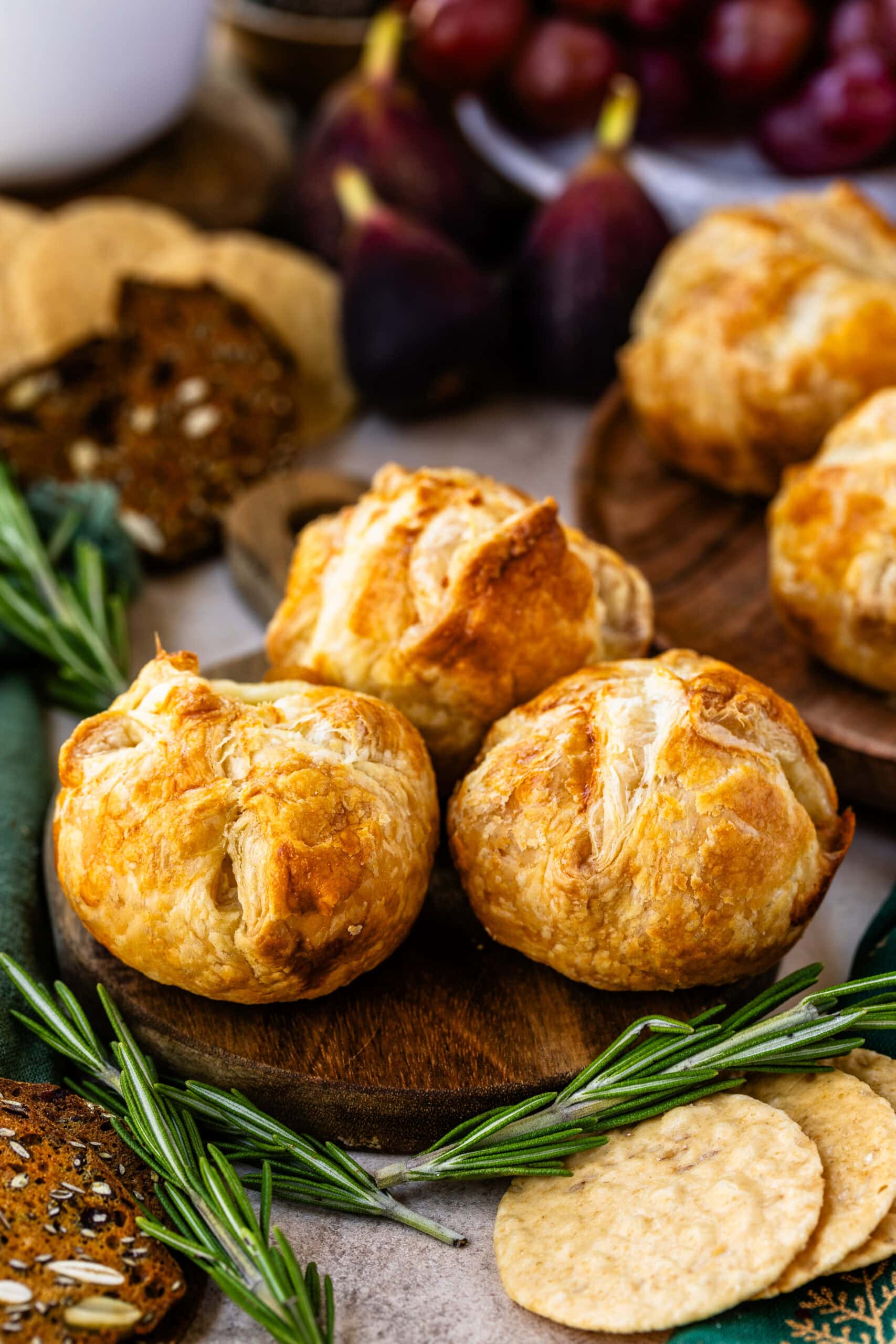 This image showcases a batch of mini baked brie pastries, golden and flaky, with beautifully crisp edges. They are arranged on a wooden board, surrounded by fresh sprigs of rosemary, seeded crackers, figs, and round flatbread crackers, adding a festive and rustic touch. The warm, golden color of the pastries creates an inviting and cozy presentation, perfect for a holiday appetizer or special gathering.