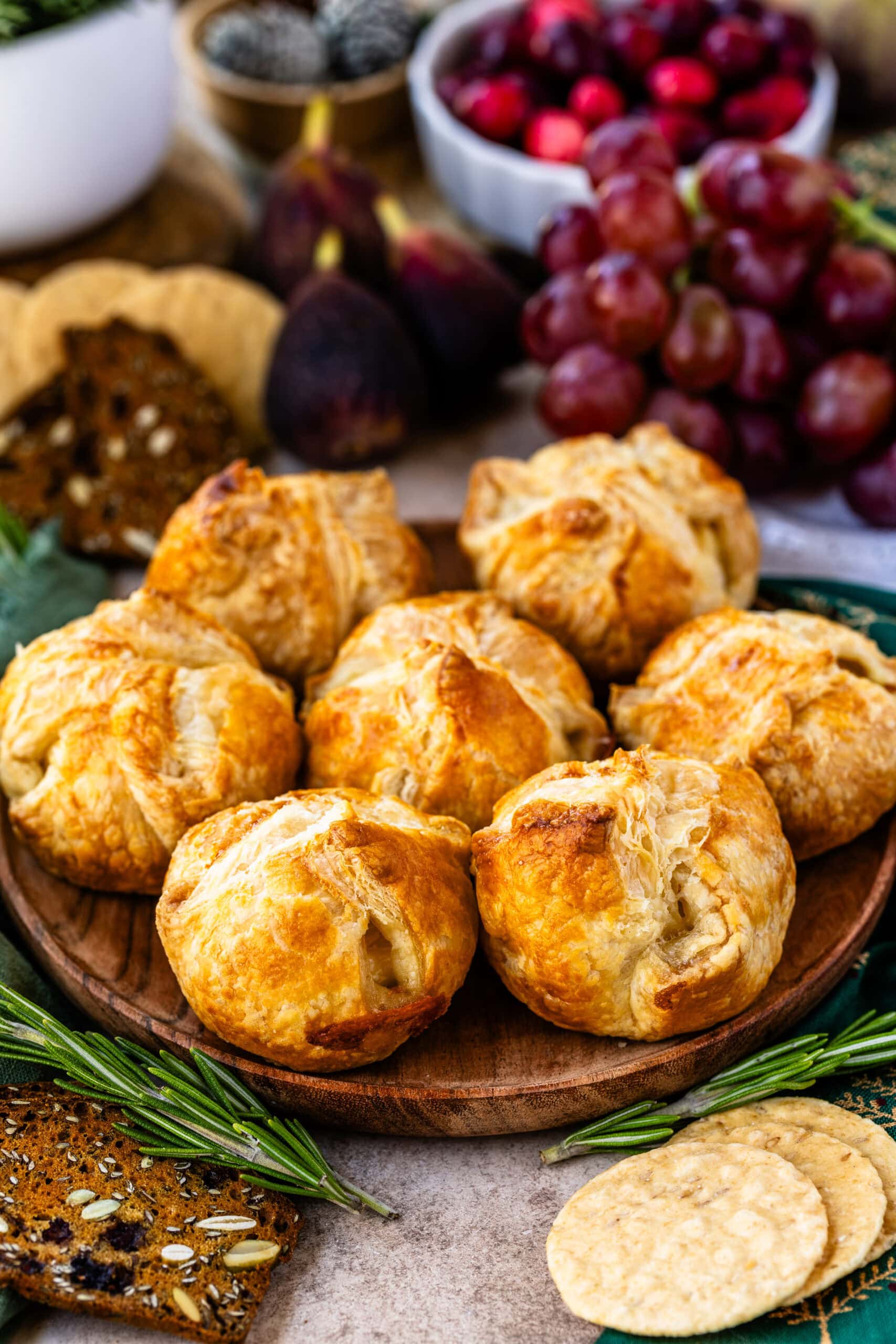 This image presents a platter of mini baked brie pastries, each golden brown with flaky, crisp layers, arranged attractively on a wooden board. The setup is surrounded by festive garnishes, including fresh figs, sprigs of rosemary, grapes, and bowls of cranberries, creating a holiday-inspired atmosphere. Seeded crackers and round flatbread crackers complete the rustic, elegant presentation, making these mini baked brie bites look deliciously inviting and perfect for a cozy gathering.