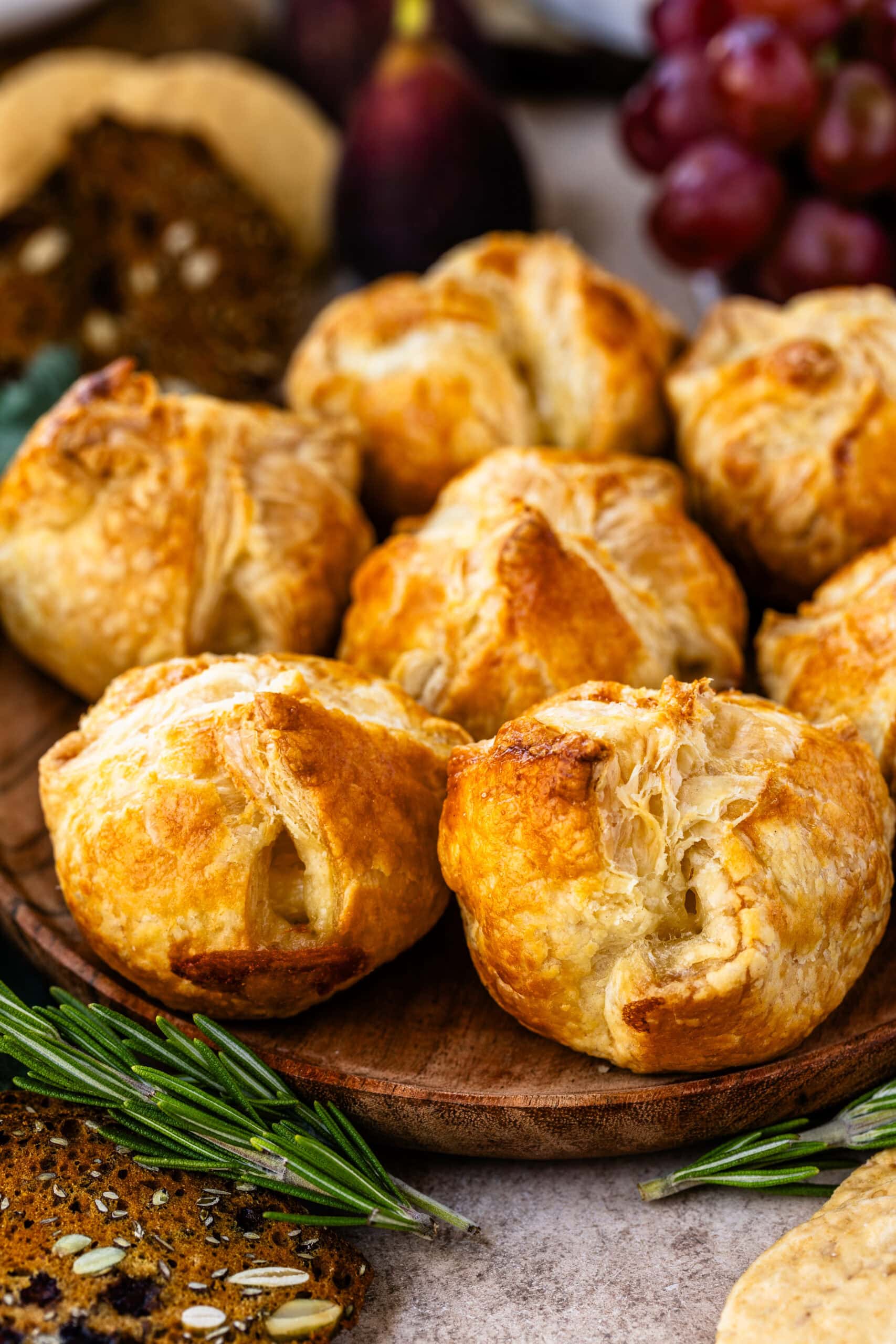 This close-up image shows a batch of mini baked brie pastries, beautifully golden and flaky, displayed on a wooden serving board. Each pastry has a crispy exterior, with delicate layers that promise a warm, gooey interior. Fresh rosemary sprigs and seeded crackers around the board enhance the rustic presentation, adding a festive touch. The setup is perfect for serving as a cozy, elegant appetizer during holiday gatherings.
