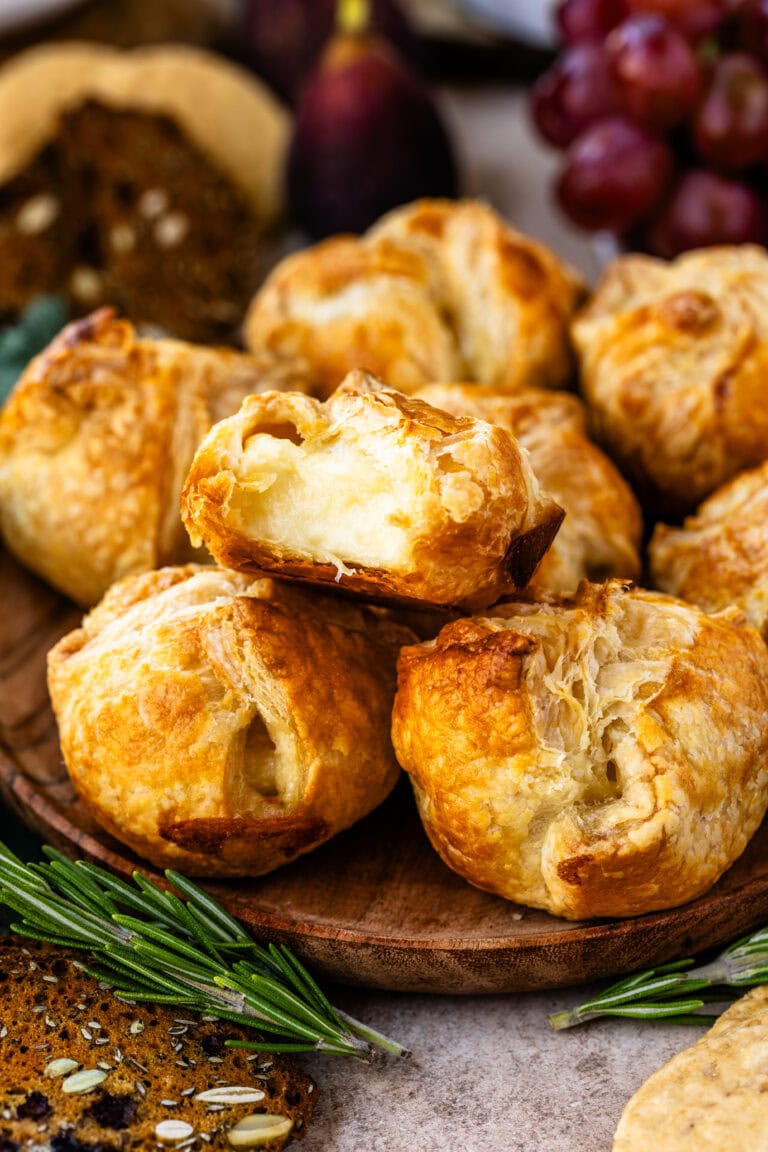 This close-up image highlights a batch of mini baked brie pastries with one pastry cut open to reveal its warm, gooey cheese filling. The golden, flaky crust contrasts beautifully with the soft, melted brie inside, making it look irresistibly delicious. Fresh rosemary sprigs and seeded crackers surround the platter, adding to the rustic and festive presentation. The setup is perfect for a holiday appetizer, inviting guests to enjoy a rich and comforting bite.