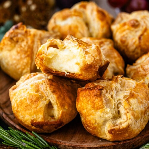 This close-up image highlights a batch of mini baked brie pastries with one pastry cut open to reveal its warm, gooey cheese filling. The golden, flaky crust contrasts beautifully with the soft, melted brie inside, making it look irresistibly delicious. Fresh rosemary sprigs and seeded crackers surround the platter, adding to the rustic and festive presentation. The setup is perfect for a holiday appetizer, inviting guests to enjoy a rich and comforting bite.