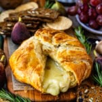 This close-up image captures a decadent baked brie with the golden, flaky pastry opened to reveal the gooey, melted cheese oozing out, creating an irresistible visual appeal. The creamy brie contrasts beautifully with the crisp pastry, making it look warm and inviting. Surrounding the brie are rustic seeded crackers and fresh sprigs of rosemary, enhancing the presentation with texture and color. The setting exudes a cozy, festive atmosphere, perfect for a holiday appetizer or a special gathering, with flavors that promise to be both rich and comforting.