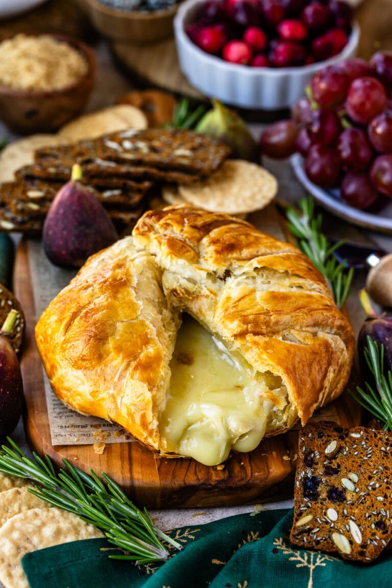 This close-up image captures a decadent baked brie with the golden, flaky pastry opened to reveal the gooey, melted cheese oozing out, creating an irresistible visual appeal. The creamy brie contrasts beautifully with the crisp pastry, making it look warm and inviting. Surrounding the brie are rustic seeded crackers and fresh sprigs of rosemary, enhancing the presentation with texture and color. The setting exudes a cozy, festive atmosphere, perfect for a holiday appetizer or a special gathering, with flavors that promise to be both rich and comforting.