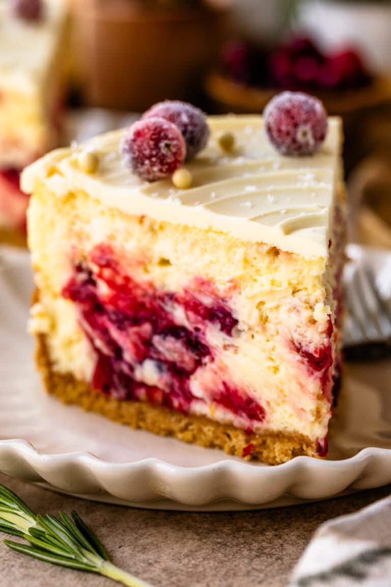 A close-up of a slice of cranberry cheesecake, highlighting the creamy texture and vibrant cranberry swirls within the filling. The top is decorated with sugared cranberries and white chocolate pearls, adding a festive touch. The slice sits on a scalloped plate, with a golden crust at the base, capturing an inviting and elegant presentation. A sprig of rosemary is placed beside the plate for a pop of green, enhancing the holiday feel.