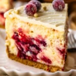 A close-up of a slice of cranberry cheesecake, highlighting the creamy texture and vibrant cranberry swirls within the filling. The top is decorated with sugared cranberries and white chocolate pearls, adding a festive touch. The slice sits on a scalloped plate, with a golden crust at the base, capturing an inviting and elegant presentation. A sprig of rosemary is placed beside the plate for a pop of green, enhancing the holiday feel.
