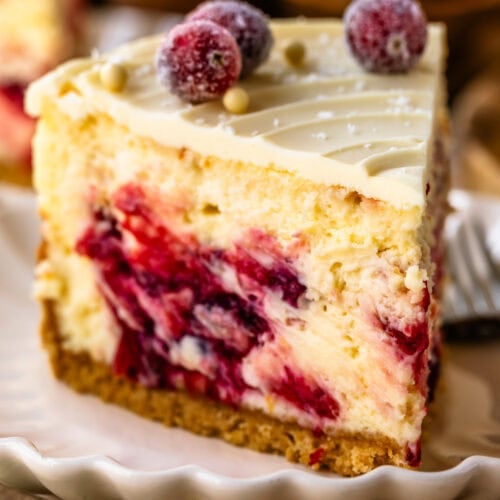 A close-up of a slice of cranberry cheesecake, highlighting the creamy texture and vibrant cranberry swirls within the filling. The top is decorated with sugared cranberries and white chocolate pearls, adding a festive touch. The slice sits on a scalloped plate, with a golden crust at the base, capturing an inviting and elegant presentation. A sprig of rosemary is placed beside the plate for a pop of green, enhancing the holiday feel.