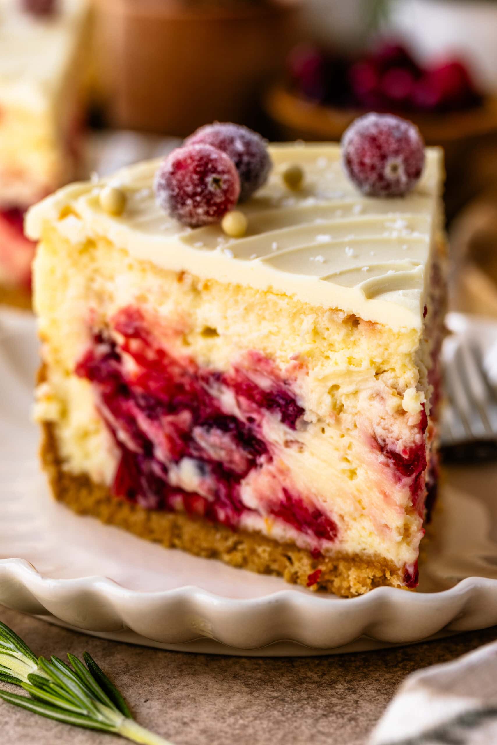 A close-up of a slice of cranberry cheesecake, highlighting the creamy texture and vibrant cranberry swirls within the filling. The top is decorated with sugared cranberries and white chocolate pearls, adding a festive touch. The slice sits on a scalloped plate, with a golden crust at the base, capturing an inviting and elegant presentation. A sprig of rosemary is placed beside the plate for a pop of green, enhancing the holiday feel.