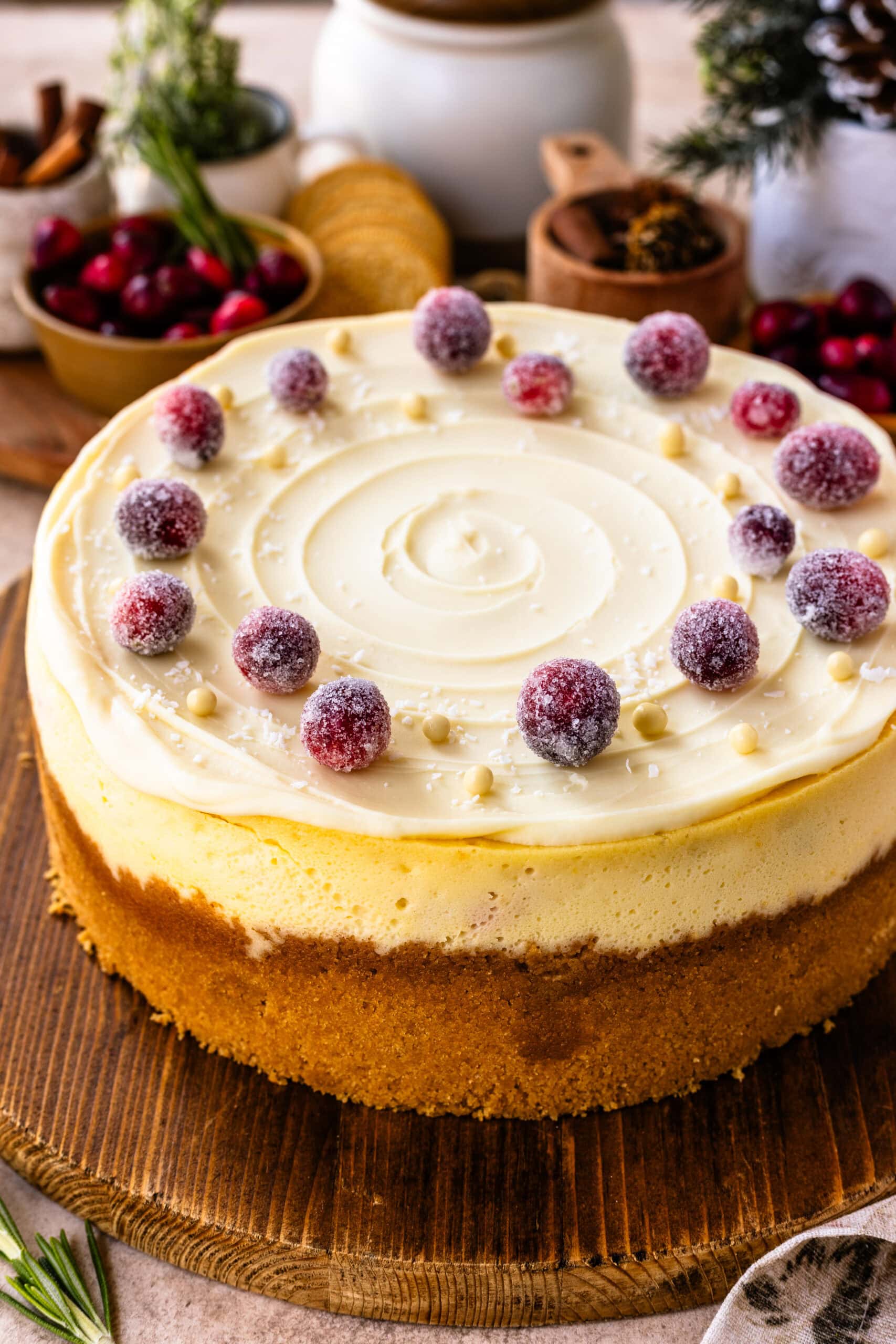 A beautifully presented whole cranberry cheesecake with a smooth cream topping, decorated with sugared cranberries and white chocolate pearls arranged around the edge. The cheesecake has a golden-brown crust and subtle swirl patterns on the creamy surface. It is placed on a rustic wooden board, with a cozy background featuring holiday-inspired decor, including fresh cranberries, greenery, and pinecones, enhancing the festive atmosphere.
