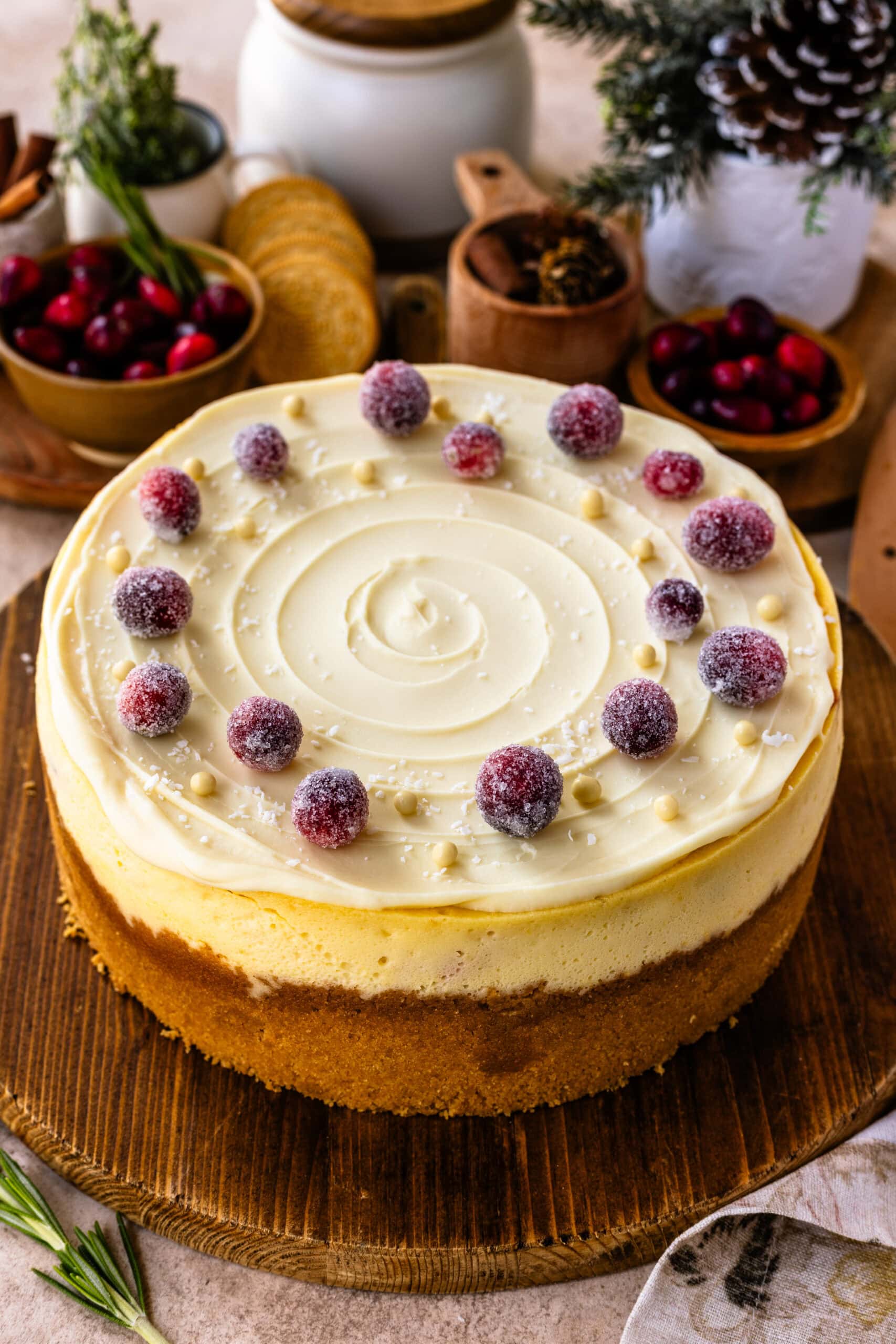 A finished cranberry cheesecake decorated with sugared cranberries and white chocolate pearls arranged around the edges, creating a festive, elegant look. The creamy, smooth topping has a subtle swirl pattern, and the cheesecake sits on a rustic wooden board. The background includes decorative elements like cranberries, cookies, and pinecones, enhancing the holiday ambiance.
