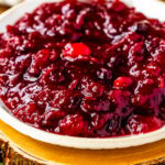 This close-up image highlights a bowl of thick, glossy cranberry sauce, placed on a rustic wooden slab that enhances the cozy, festive feel. The rich red color and chunky texture of the cranberry sauce are visually striking, with whole cranberries adding depth and texture. Fresh rosemary sprigs are arranged nearby, contributing a touch of greenery to the setup. The cranberry sauce looks enticing and perfect for a holiday meal, with the rustic background elements creating a warm, inviting presentation.
