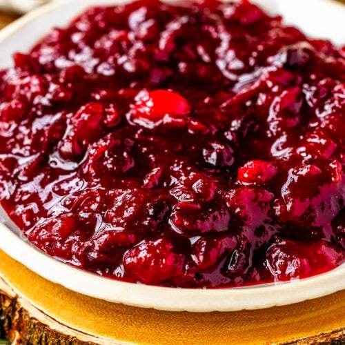This close-up image highlights a bowl of thick, glossy cranberry sauce, placed on a rustic wooden slab that enhances the cozy, festive feel. The rich red color and chunky texture of the cranberry sauce are visually striking, with whole cranberries adding depth and texture. Fresh rosemary sprigs are arranged nearby, contributing a touch of greenery to the setup. The cranberry sauce looks enticing and perfect for a holiday meal, with the rustic background elements creating a warm, inviting presentation.