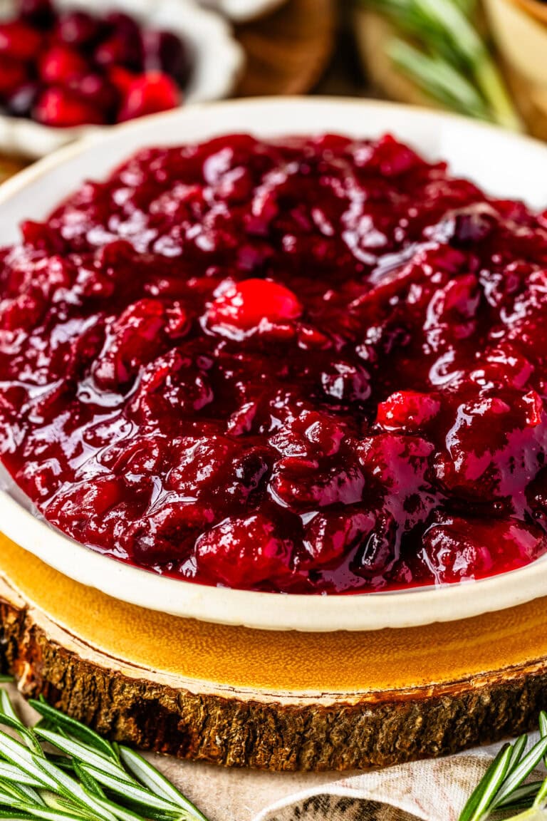 This close-up image highlights a bowl of thick, glossy cranberry sauce, placed on a rustic wooden slab that enhances the cozy, festive feel. The rich red color and chunky texture of the cranberry sauce are visually striking, with whole cranberries adding depth and texture. Fresh rosemary sprigs are arranged nearby, contributing a touch of greenery to the setup. The cranberry sauce looks enticing and perfect for a holiday meal, with the rustic background elements creating a warm, inviting presentation.