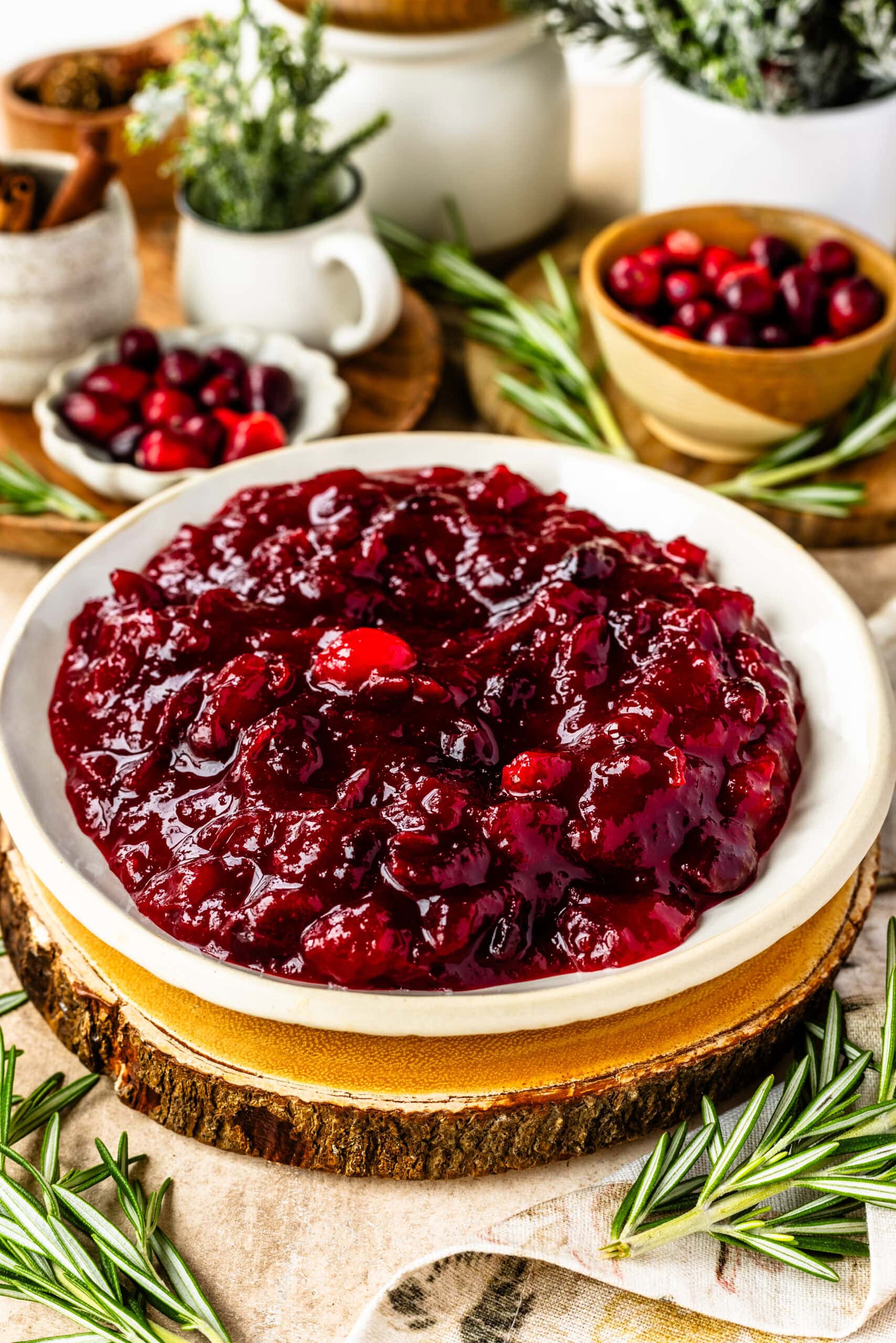 This image presents a beautifully styled bowl of cranberry sauce, thick and glossy with a deep red hue, placed on a rustic wooden slab that adds warmth to the display. The cranberry sauce looks chunky, with whole cranberries visible, inviting a rich and homemade texture. Surrounding the setup are sprigs of fresh rosemary and small bowls of cranberries in the background, enhancing the festive, holiday atmosphere. The combination of natural textures and vibrant colors creates an appealing and cozy presentation, perfect for a seasonal meal.
