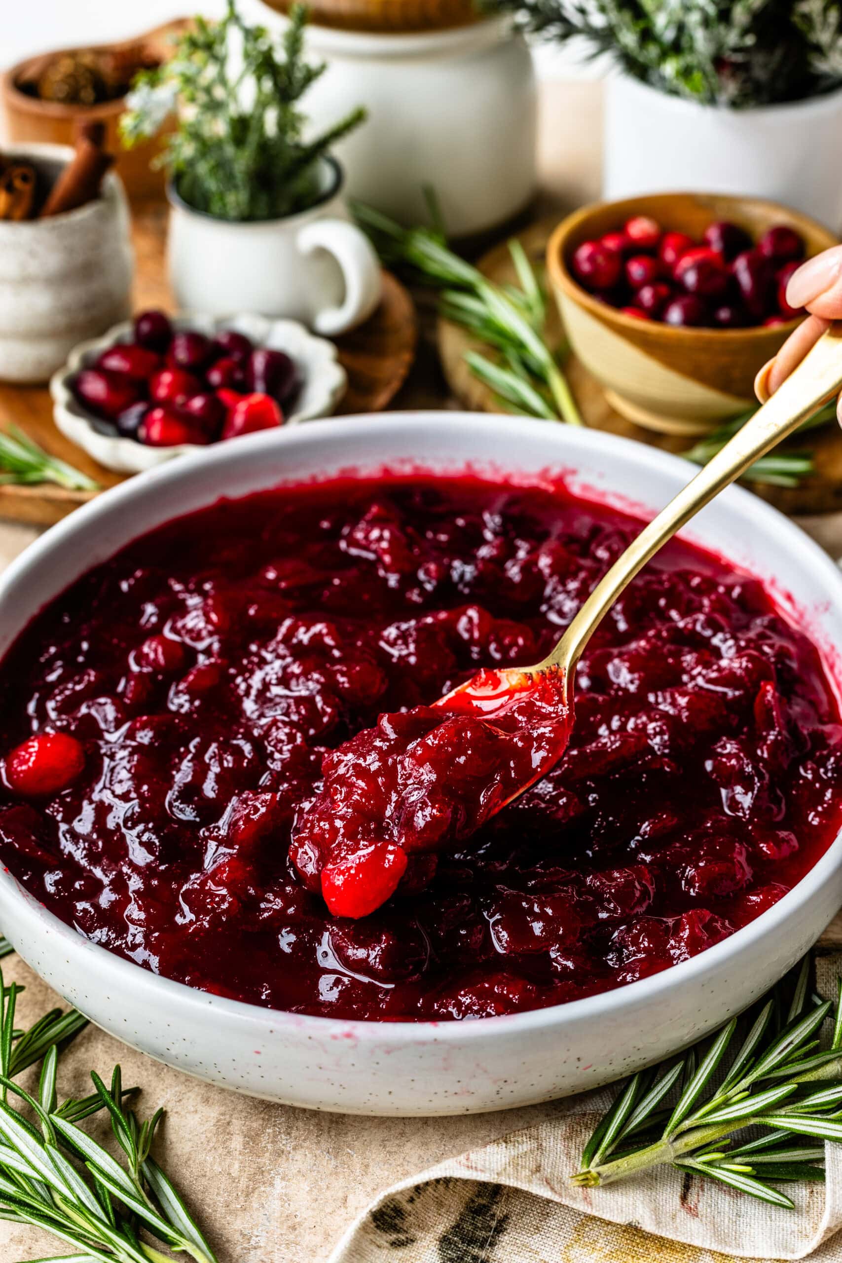 The image captures a bowl of glossy, vibrant cranberry sauce with a thick and luscious texture, enhanced by whole cranberries and a rich red color. A golden spoon, held by a hand, is scooping a generous portion of the sauce, showcasing its thick consistency. Fresh rosemary sprigs are placed around the bowl, while small dishes with extra cranberries and cinnamon sticks are visible in the softly focused background, creating a cozy and festive ambiance. The composition beautifully highlights the holiday seasonโs warmth and flavor, making the cranberry sauce look delicious and inviting.